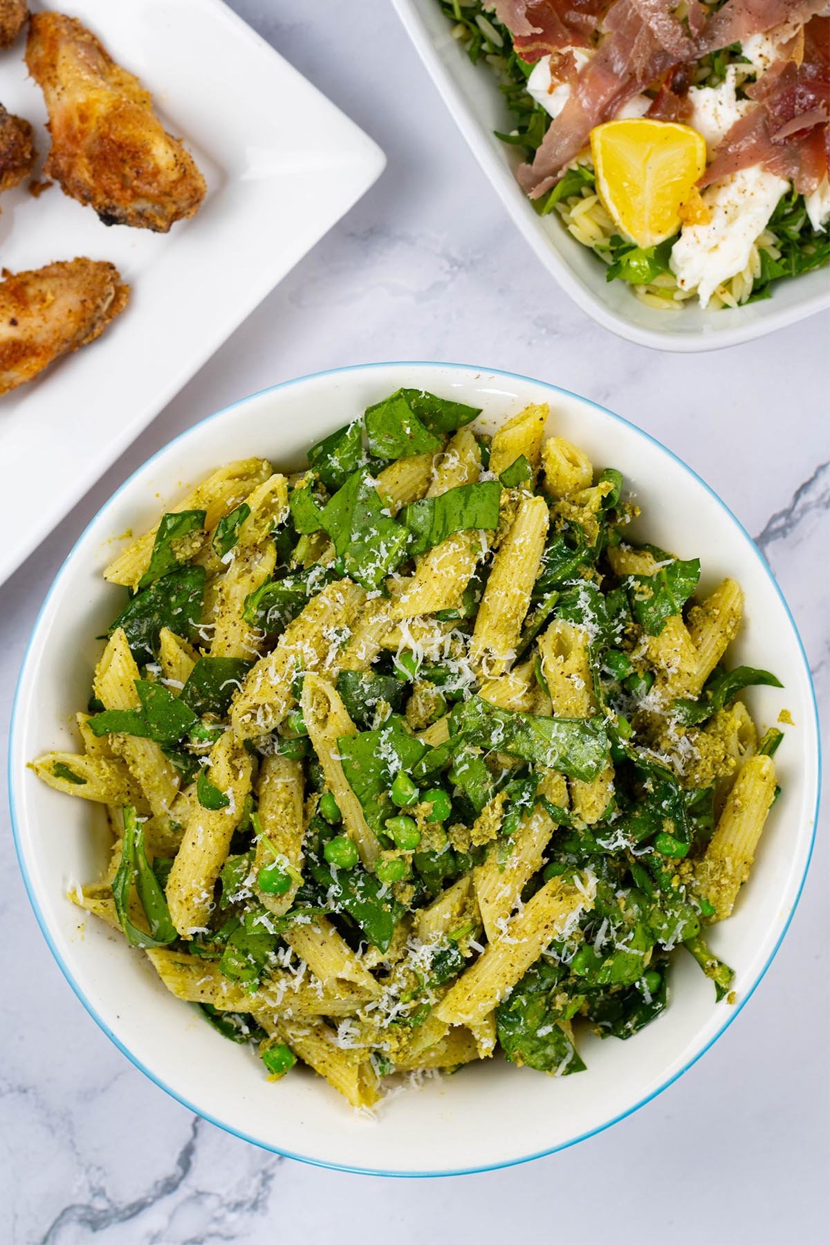 Pesto, peas and spinach pasta salad in a light blue trimmed serving bowl with a platter of cajun wings and dish of crispy prosciutto and orzo salad in the background.