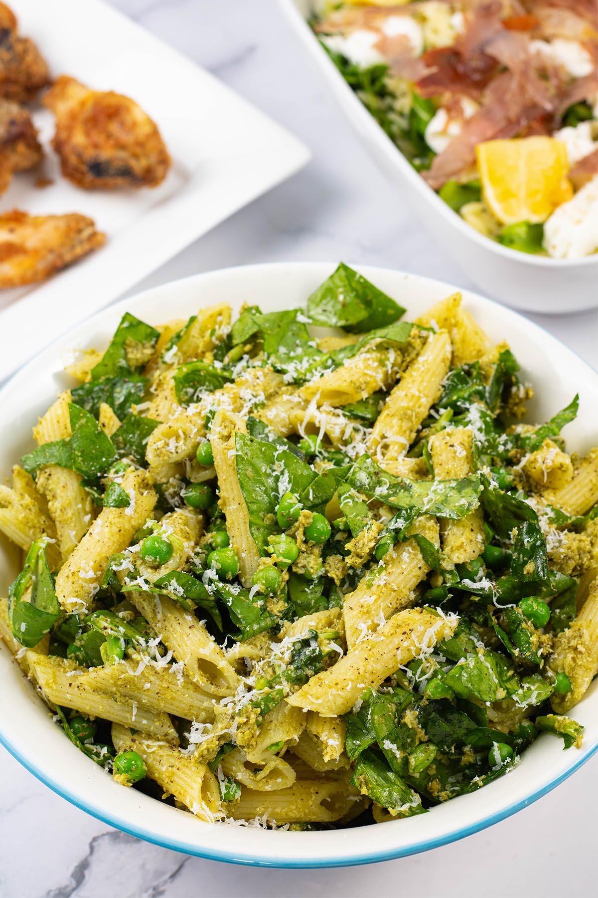 Pesto, peas and spinach pasta salad in a light blue trimmed serving bowl with a platter of cajun wings and dish of crispy prosciutto and orzo salad in the background.