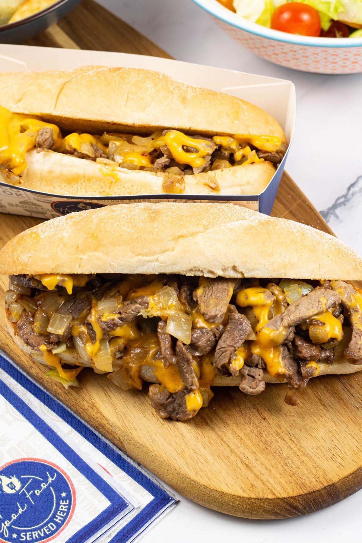 Two Philly cheesesteaks on a wooden board with a bowl of salad in the background.