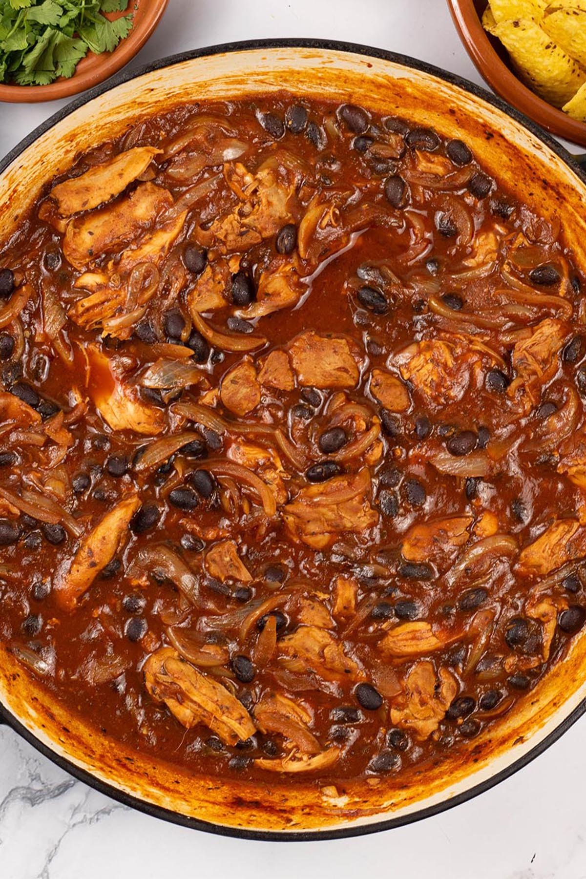 Pulled chicken and black bean chilli in a large round shallow casserole dish with dishes of coriander and tortilla crisps on the side.