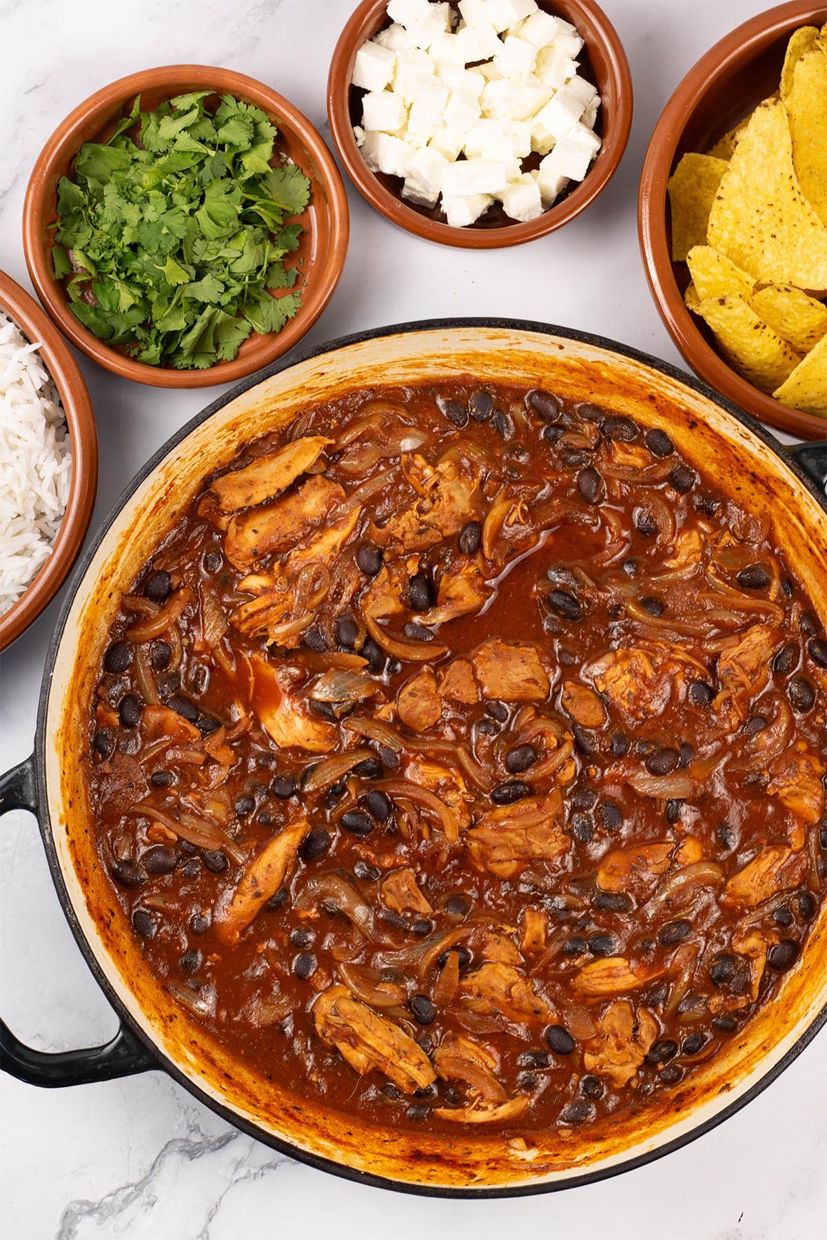 Pulled chicken and black bean chilli in a large round shallow casserole dish with dishes of rice, coriander, feta cheese and tortilla crisps on the side.