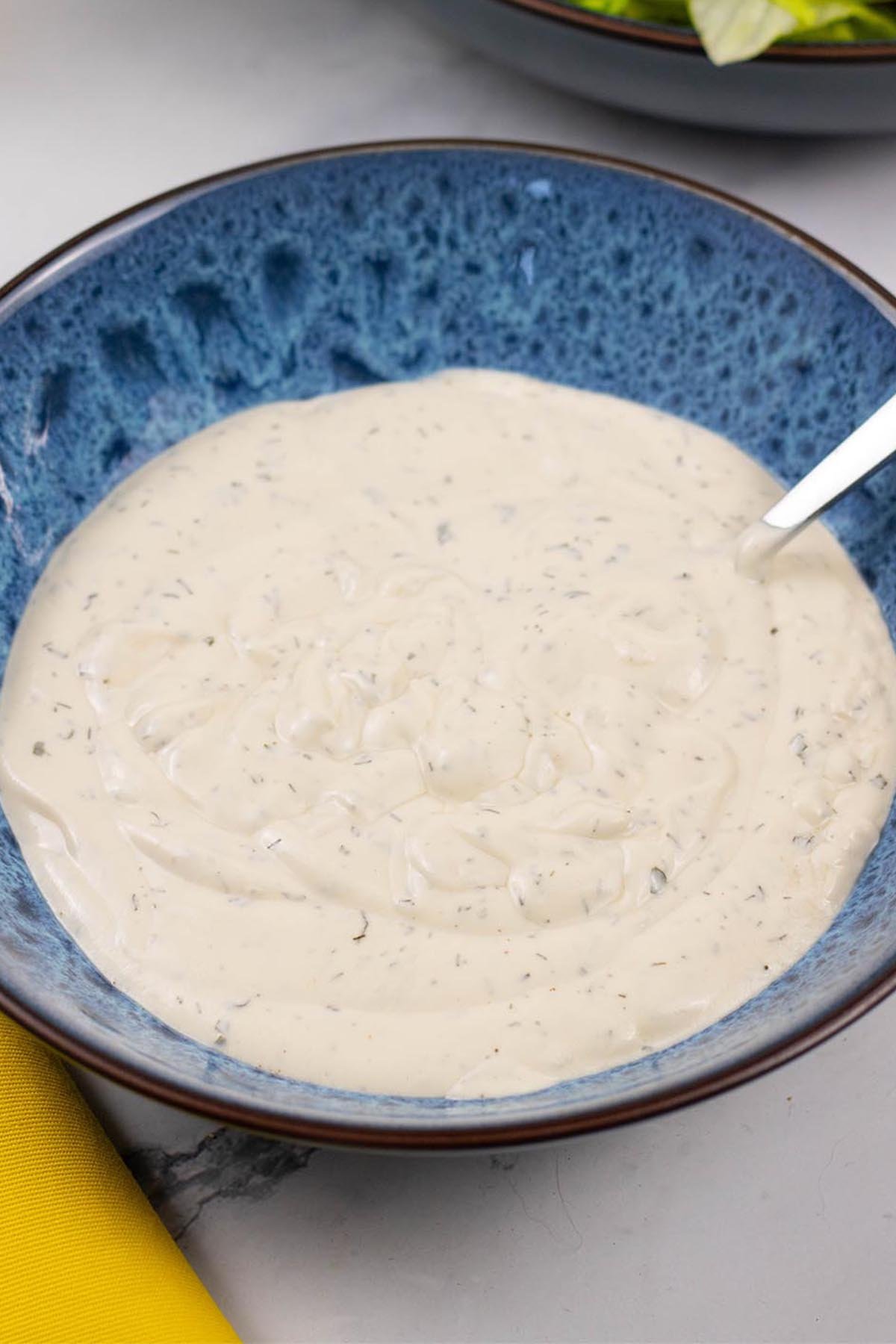Ranch dressing in a blue patterned serving bowl with spoon and a bowl of lettuce salad in the background.