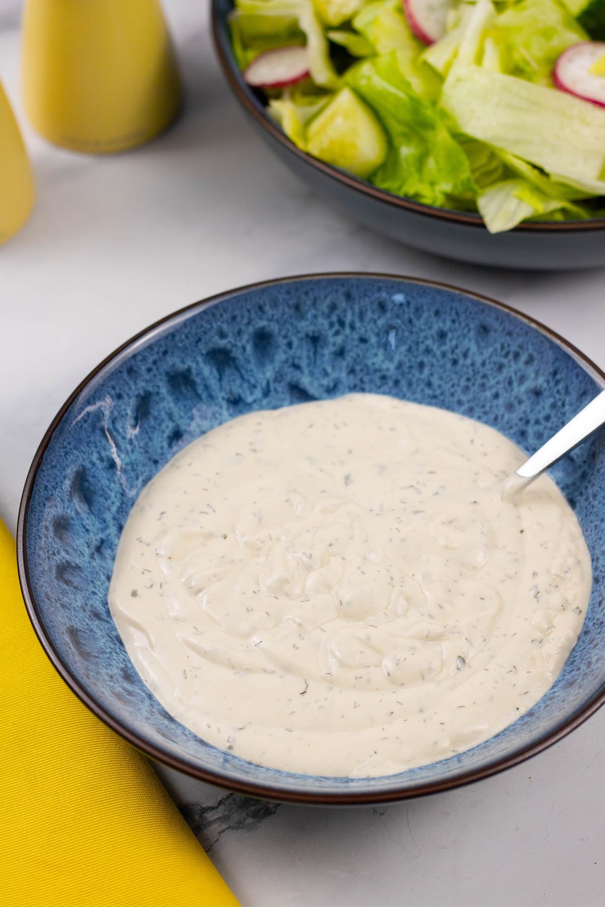 Ranch dressing in a blue patterned serving bowl with spoon and a bowl of lettuce salad in the background.