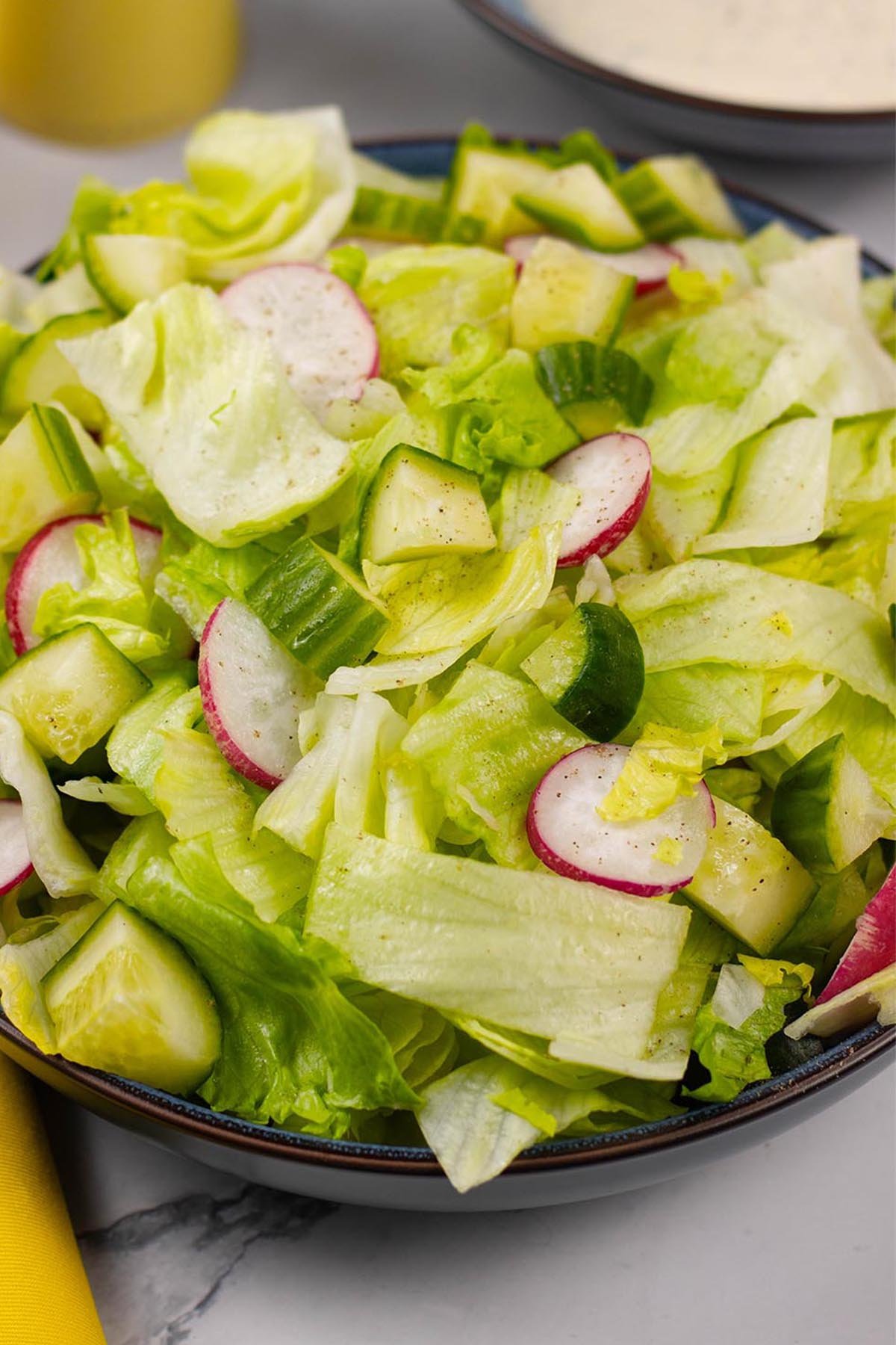 Close up of simple crunchy lettuce salad in a blue serving bowl with a small bowl of ranch dressing and yellow salt and pepper grinders in the background.
