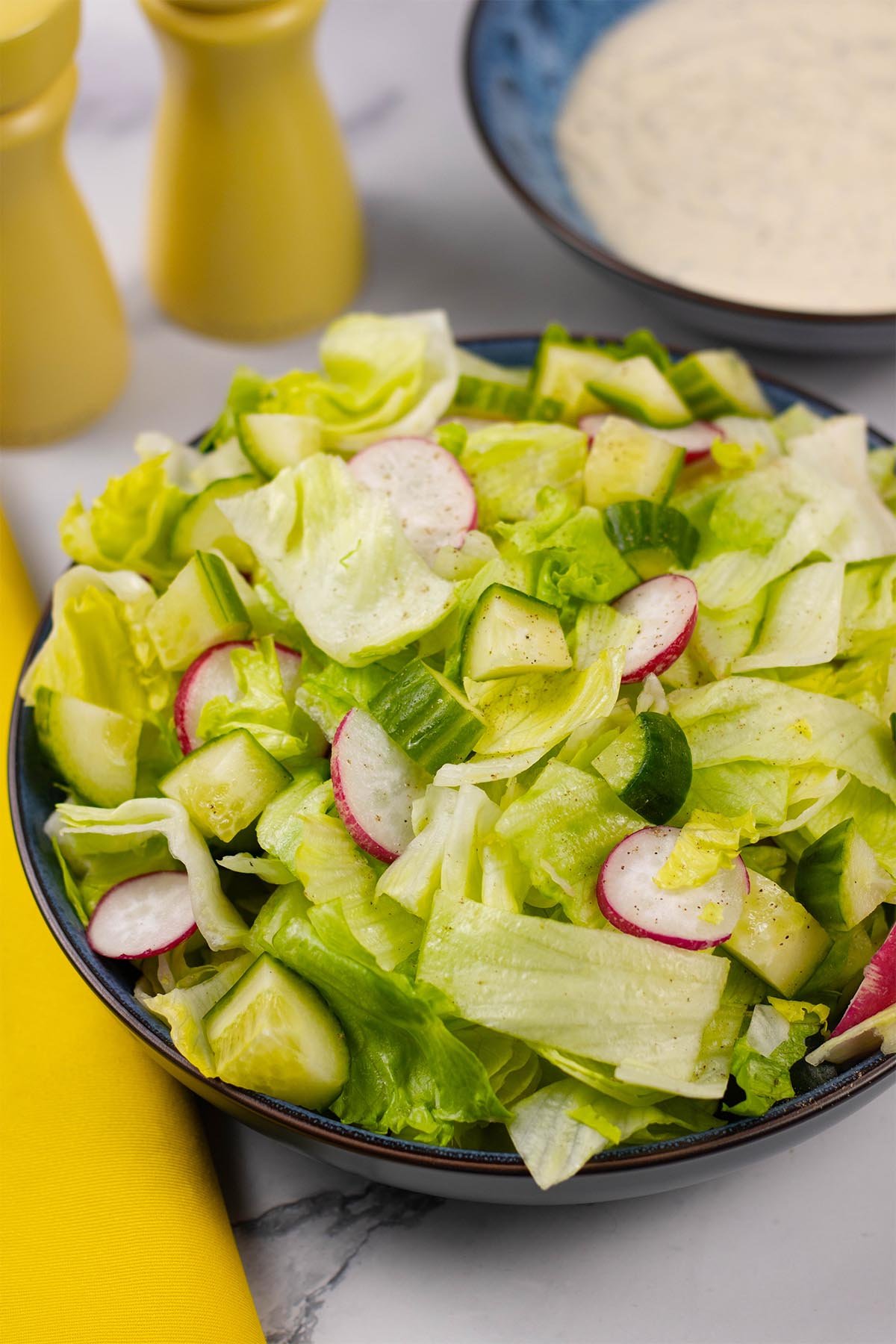 Simple crunchy lettuce salad in a blue serving bowl with a small bowl of ranch dressing and yellow salt and pepper grinders in the background.