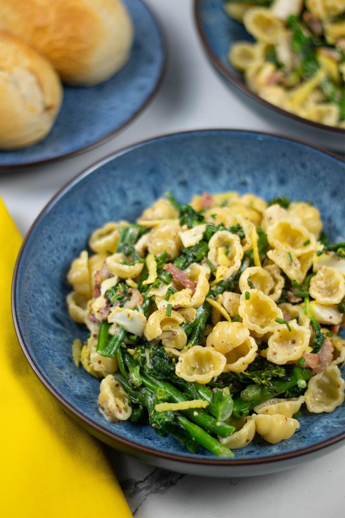 Two portions of smoked haddock with tenderstem broccoli, spinach and conchiglie in blue patterned bowls with a plate of crusty bread on the side.