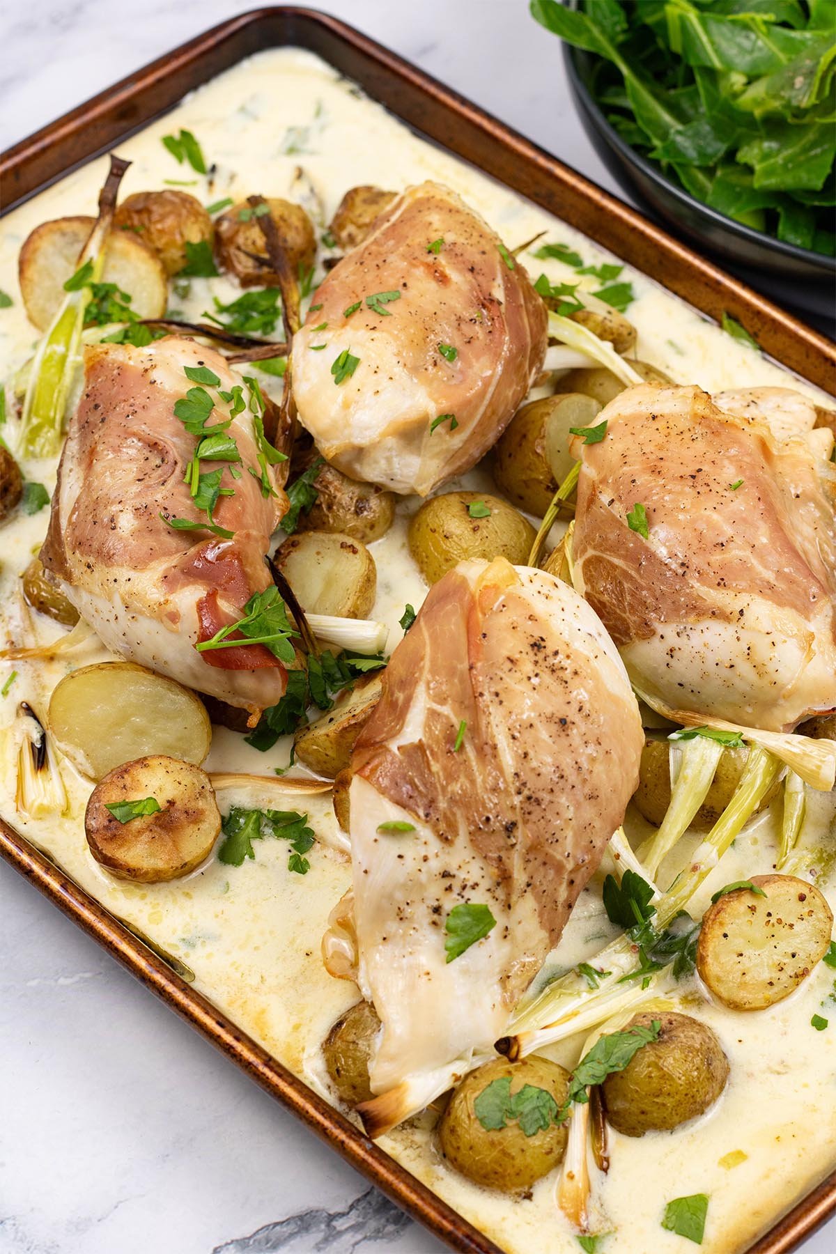 Four portions of parma ham, chicken and creamy potato traybake on a large baking tray, with a bowl of spring greens the side.