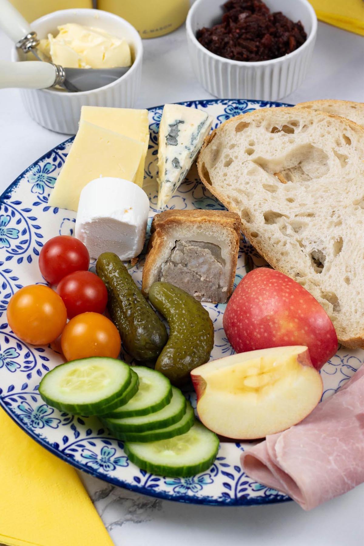 A portion of ploughman's platter on a blue and white floral patterned dinner plate with small dishes of butter and chutney on the side.