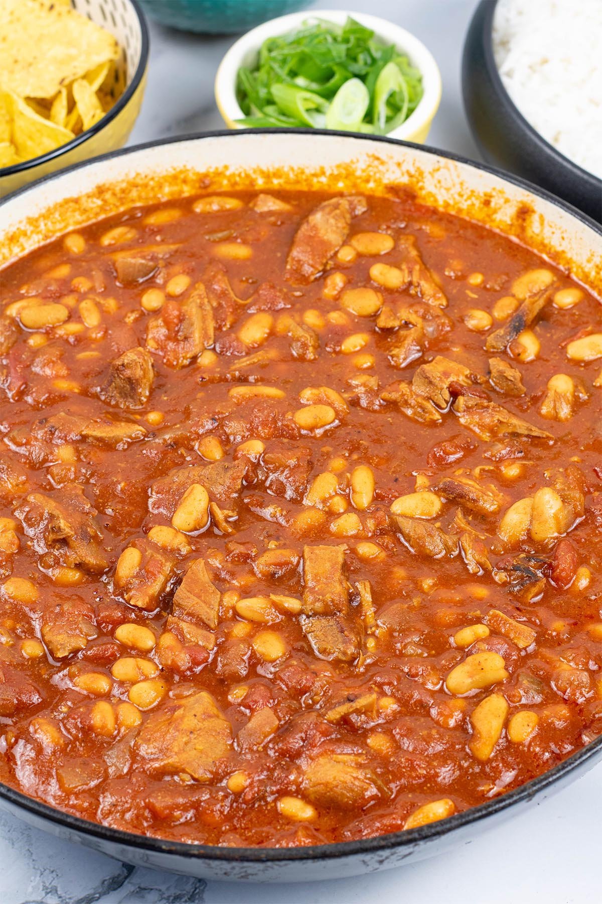 Slow cooked pork shoulder chilli in a large round casserole with bowls of nachos, spring onions and rice in the background.