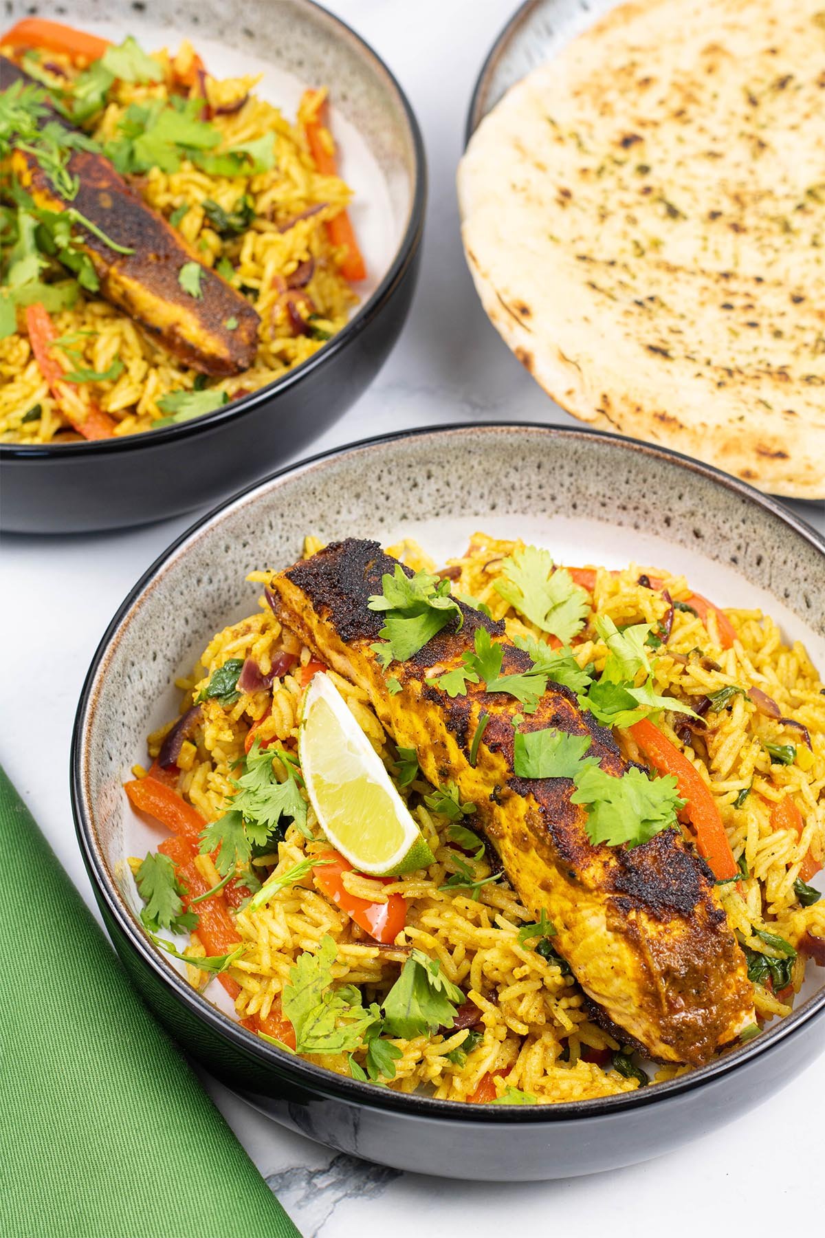 2 portions of Balti salmon rice bowls in black and white bowls with a wedge of lime and plate of garlic bread on the side.