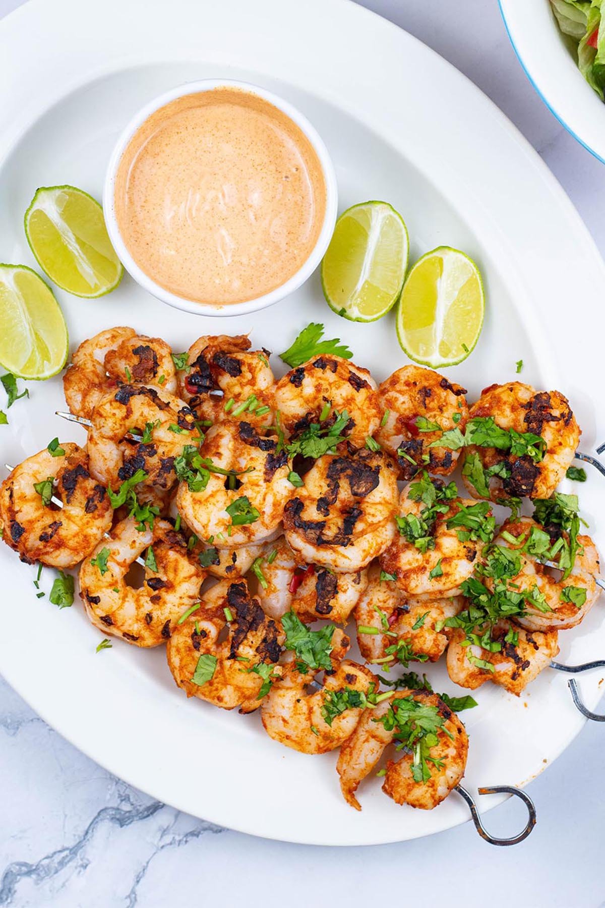 Chilli and lime prawn skewers garnished with chopped coriander on a white oval platter with lime wedges and a dish of spicy mayo.