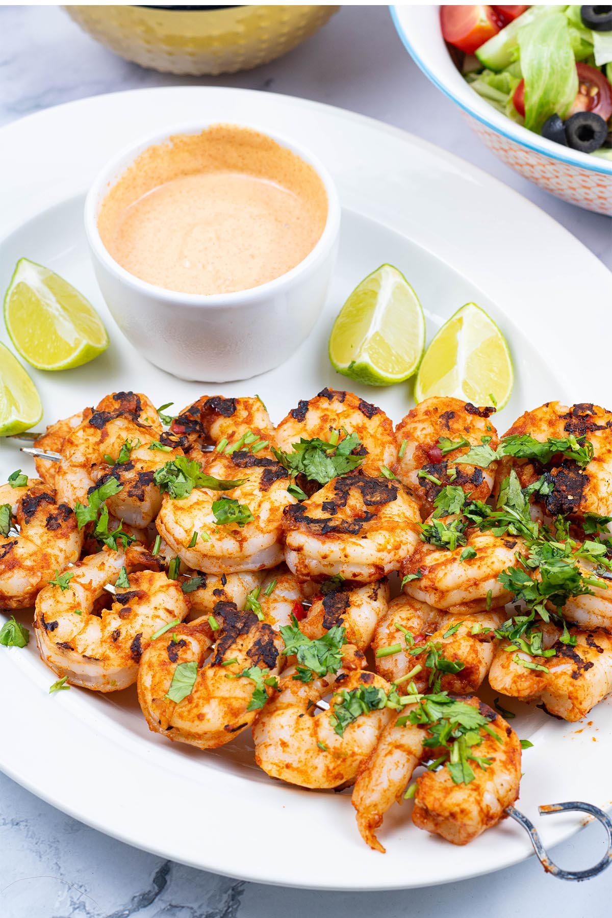 Chilli and lime prawn skewers garnished with chopped coriander on a white oval platter with lime wedges and a dish of spicy mayo.