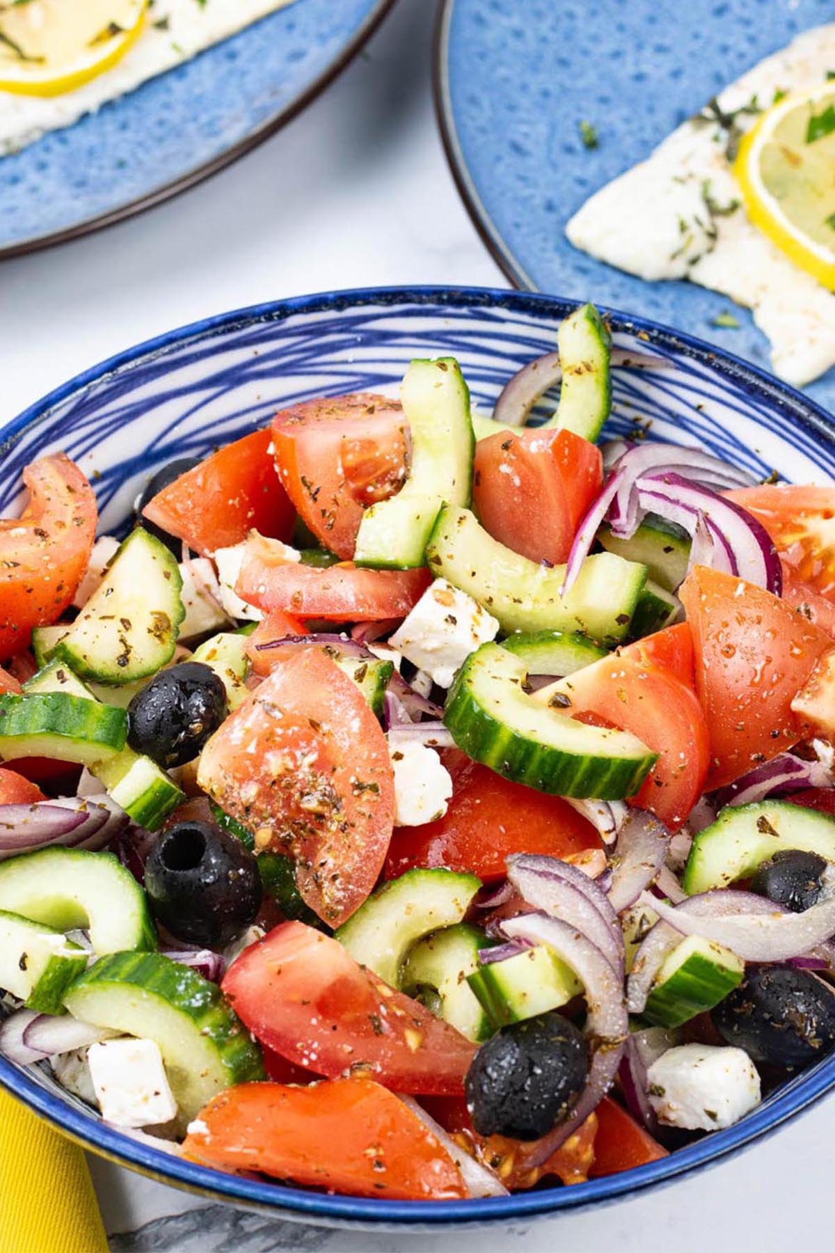 Greek salad in a blue and white striped bowl with plates of oregano and lemon barbecued sea bass in the background.