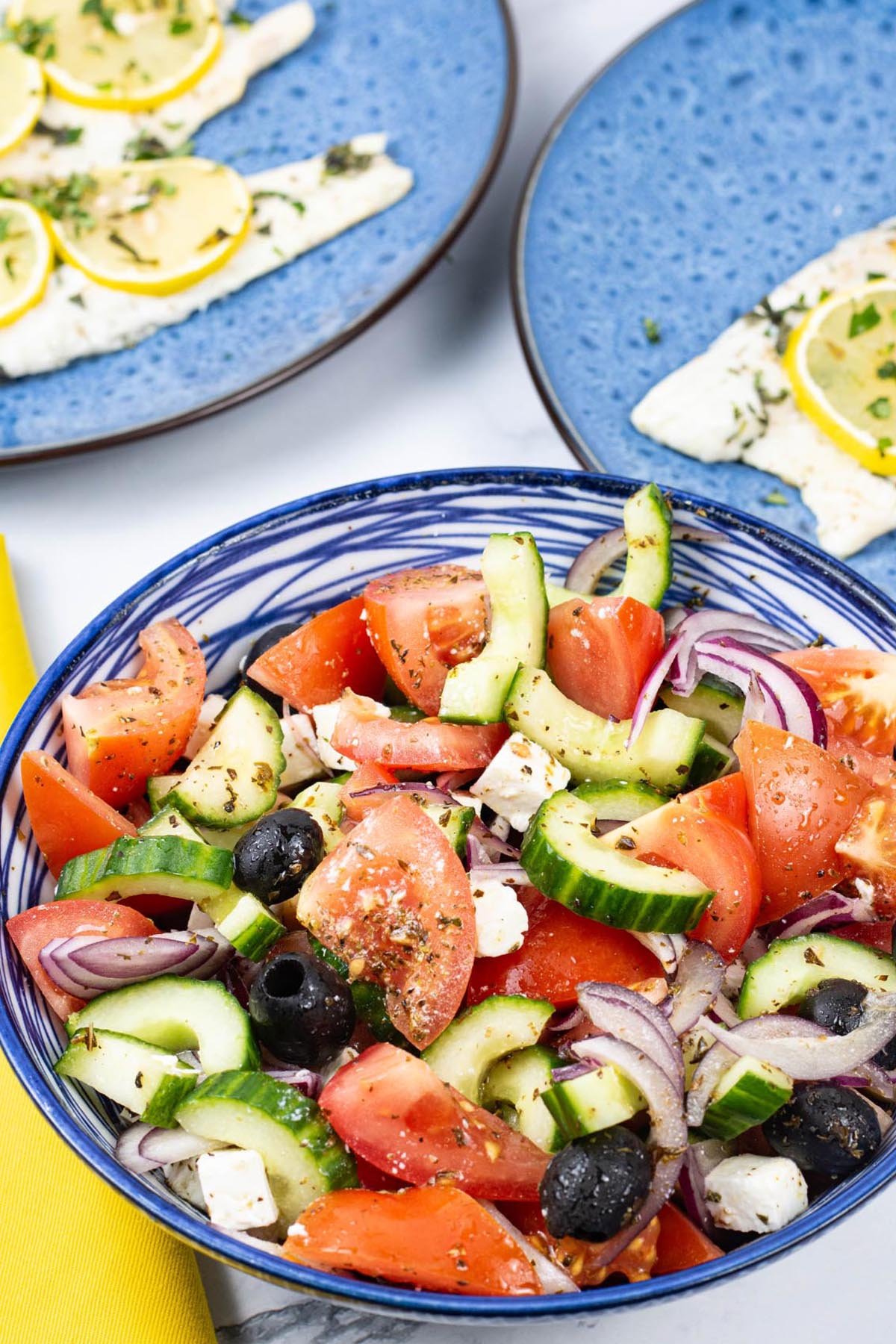 Greek salad in a blue and white striped bowl with plates of oregano and lemon barbecued sea bass in the background.