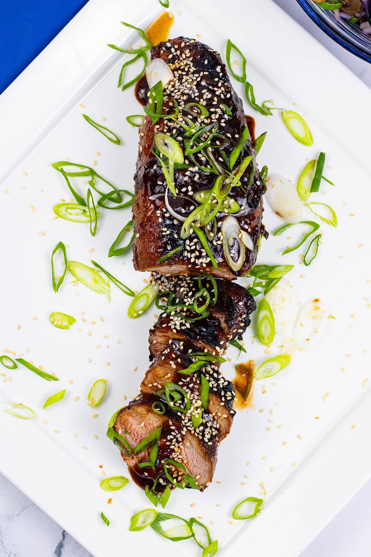 Korean barbecued pork fillet sliced and garnished with spring onions on a square white platter.