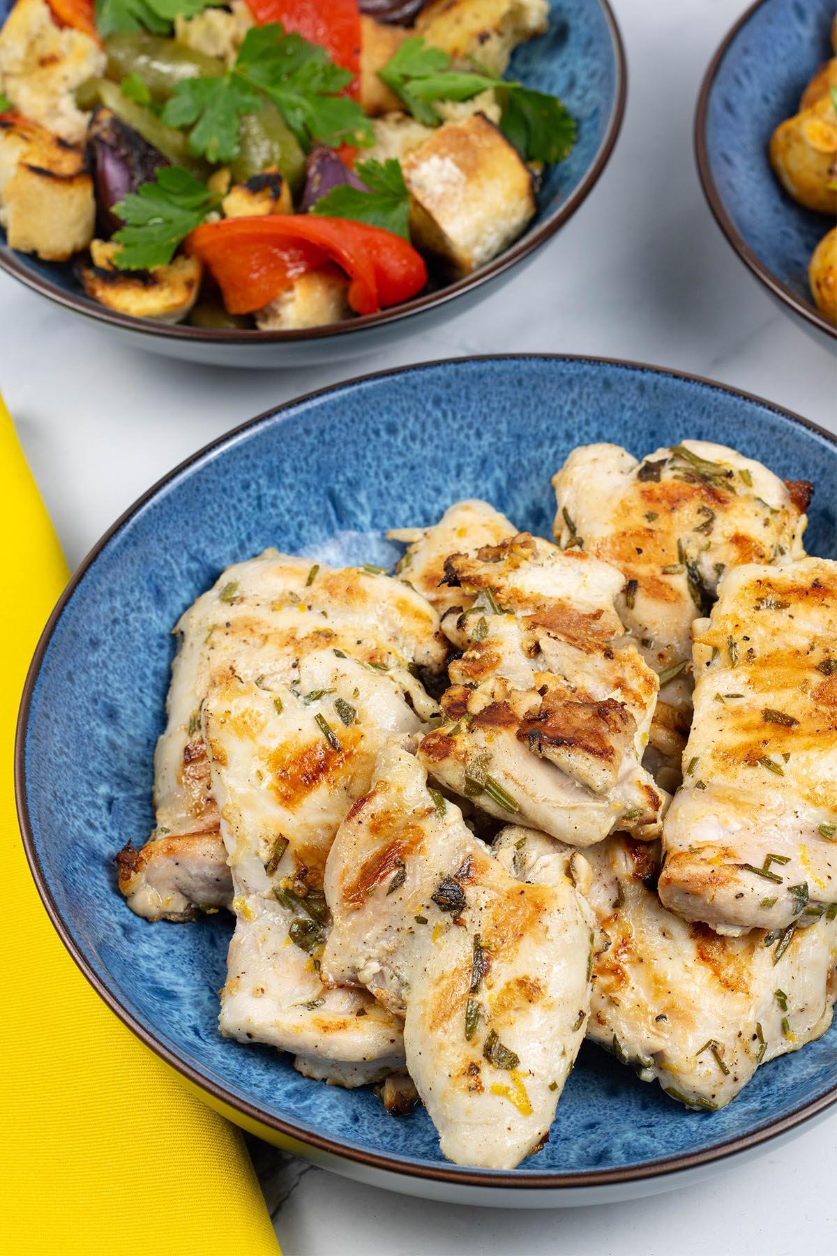 Lemon herb chicken thighs in a blue patterned serving bowl with a dish of barbecued mixed pepper salad on the side.