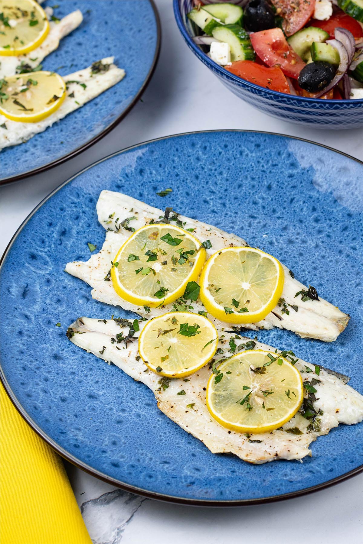 2 x oregano and lemon barbecued sea bass fillets on a blue dinner plate with a bowl of Greek salad on the side.