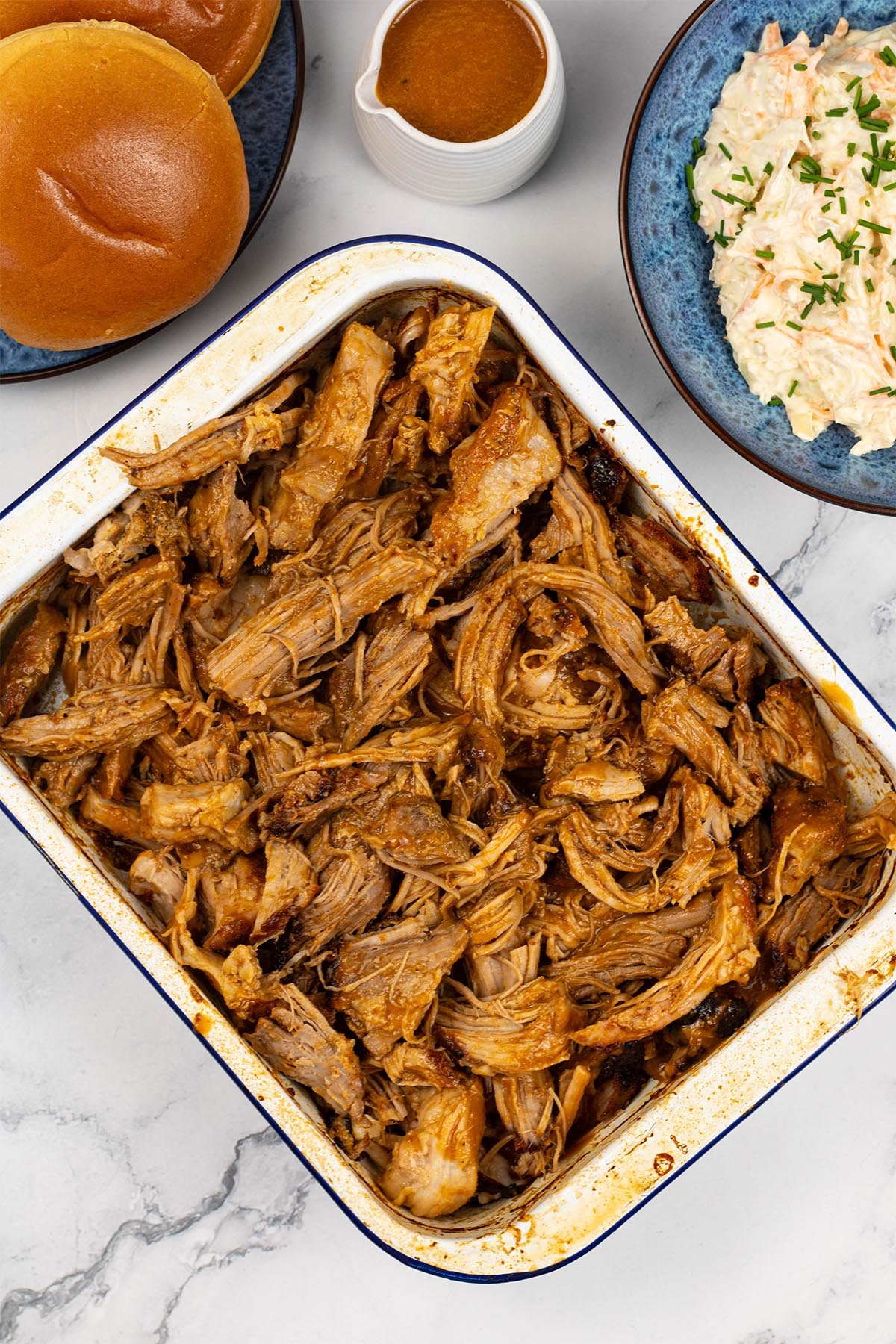 BBQ pulled pork in a blue rimmed white rectangle enamel roasting tin, with a jug of BBQ sauce, bowl of coleslaw and brioche buns in the background.