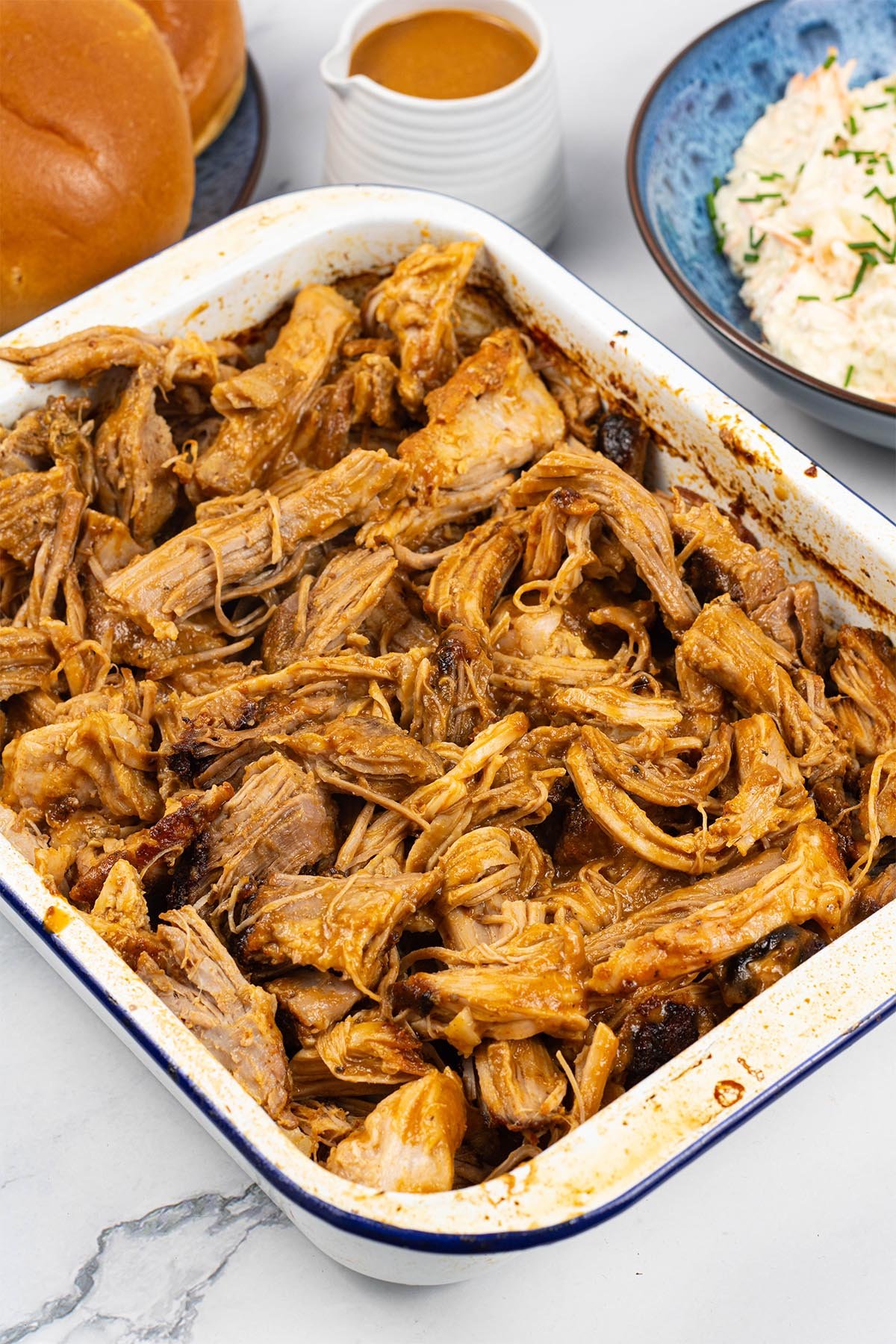 BBQ pulled pork in a blue rimmed white rectangle enamel roasting tin, with a jug of BBQ sauce, bowl of coleslaw and brioche buns in the background.