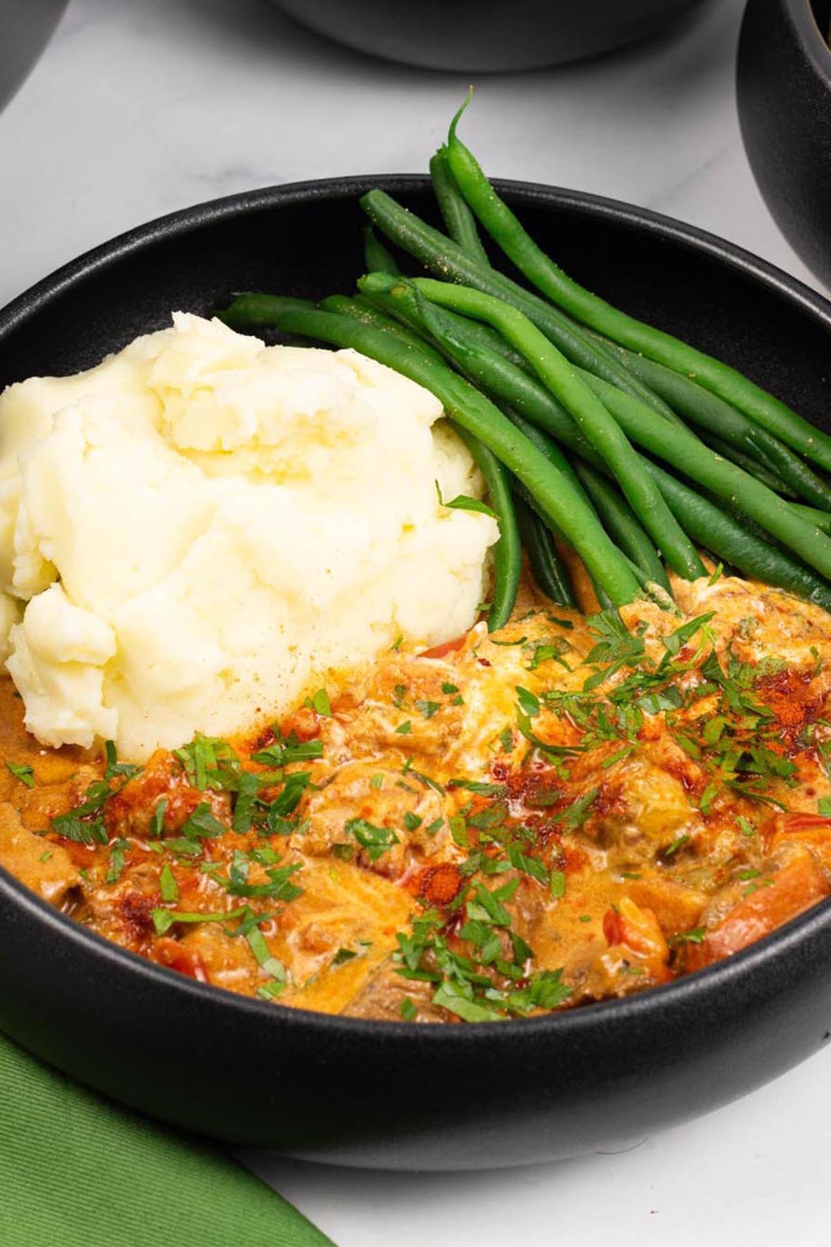 A portion of slow cooker beef goulash in a black bowl with mashed potatoes and green beans, and a green napkin on the side.