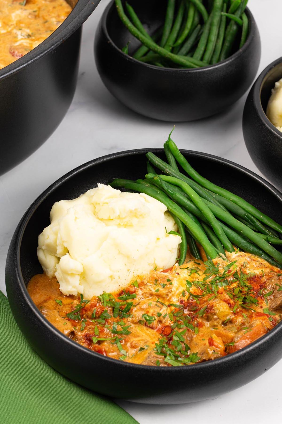 A portion of slow cooker beef goulash in a black bowl with mashed potatoes and green beans, and a green napkin on the side.
