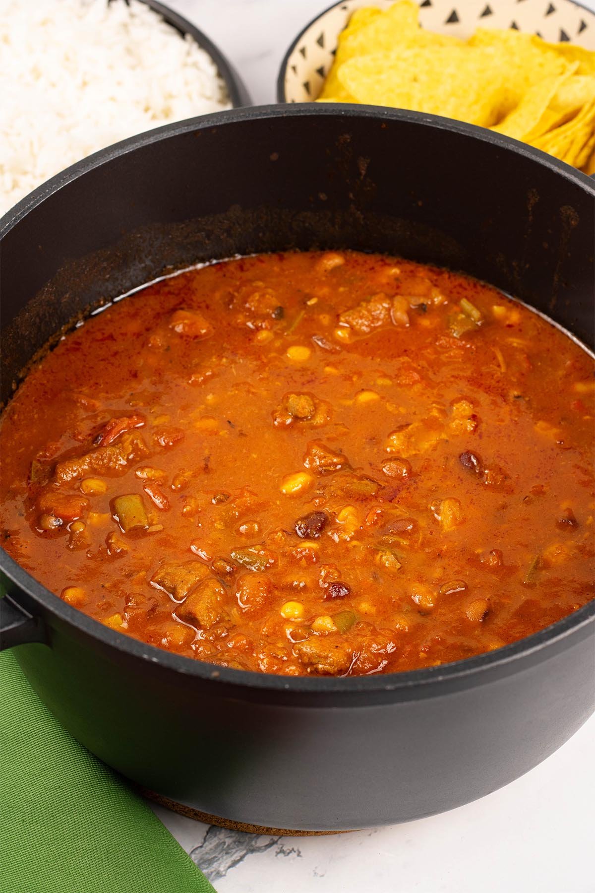 Smoky pork and beans made in the Thermomix in a black casserole pot, with serving bowls of rice and nachos in the background.