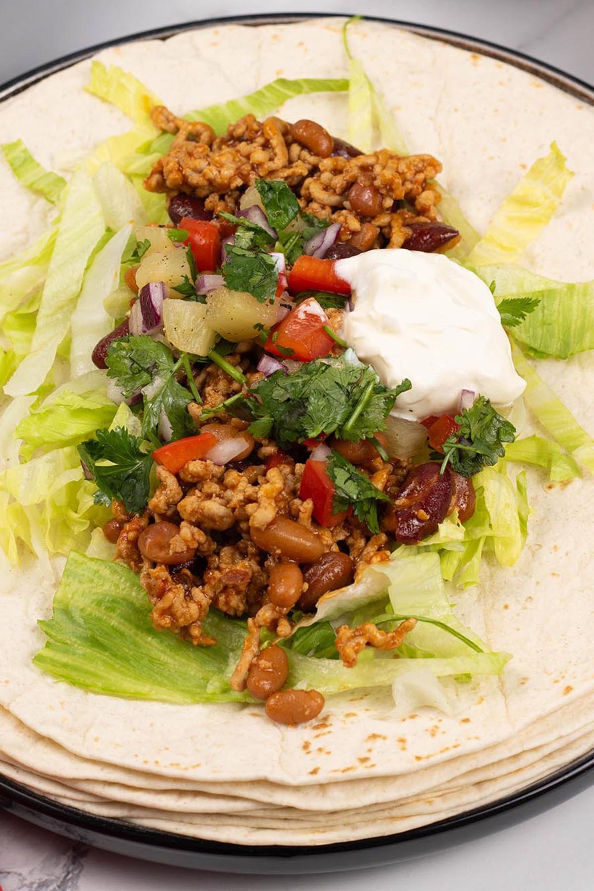 A smoky pork tortilla sitting on a stack of soft tortilla wraps, garnished with pineapple salsa and sour cream.