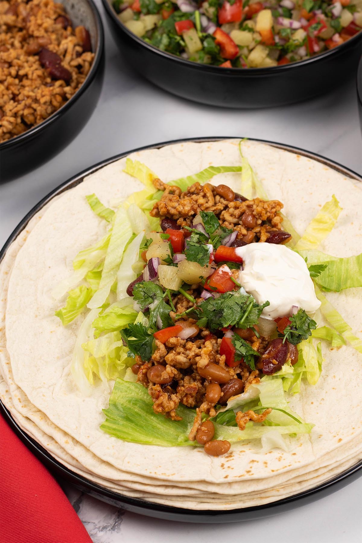 A smoky pork tortilla sitting on a stack of soft tortilla wraps, garnished with pineapple salsa and sour cream.
