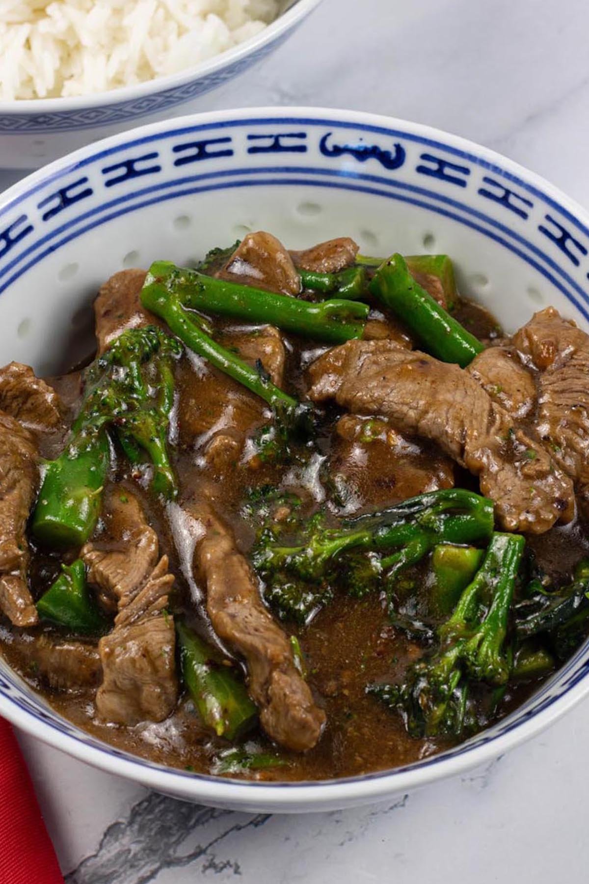 Spiced beef and broccoli in a blue and white oriental bowl with a bowl of steamed jasmine rice in the background.