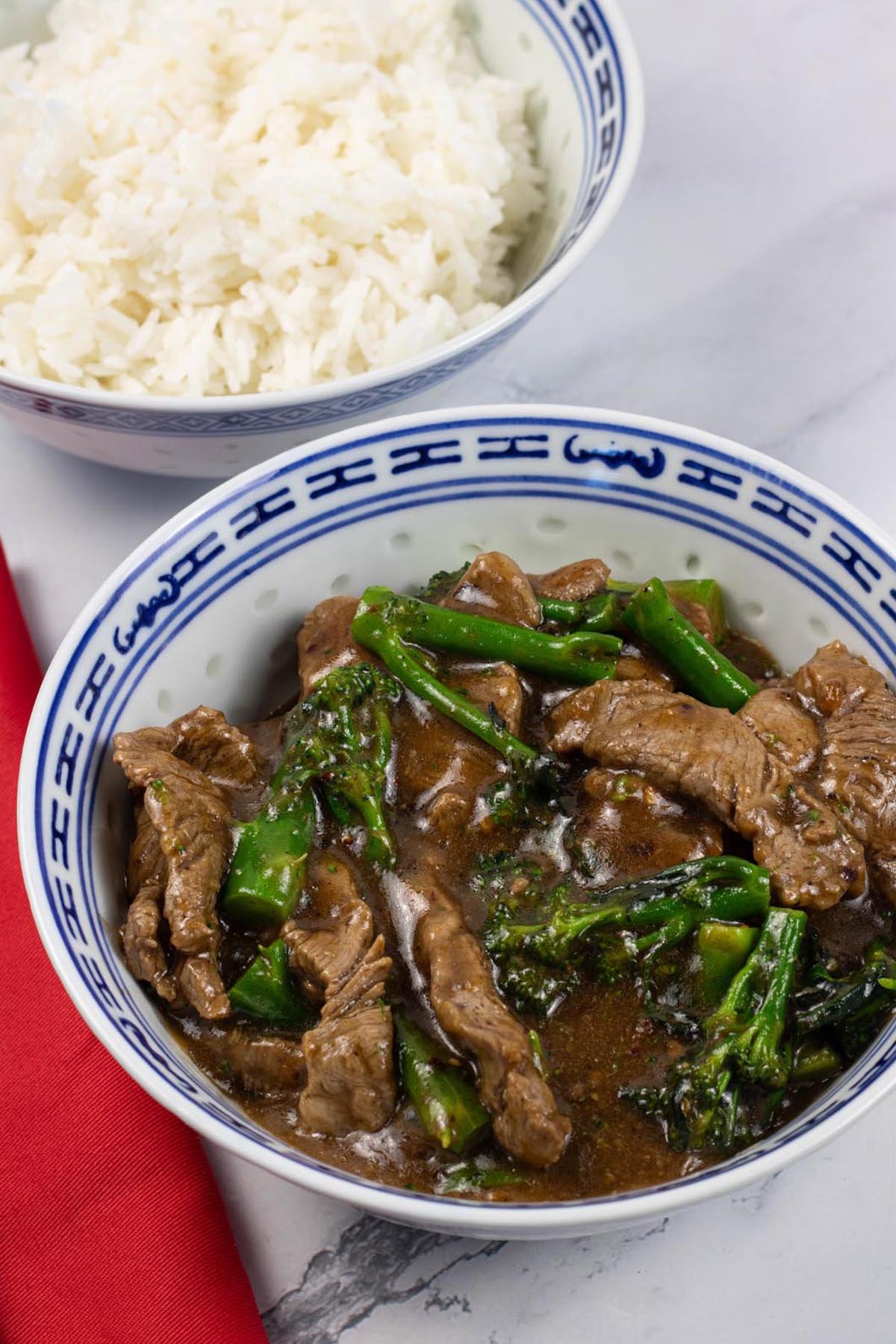Spiced beef and broccoli in a blue and white oriental bowl with a bowl of steamed jasmine rice in the background.