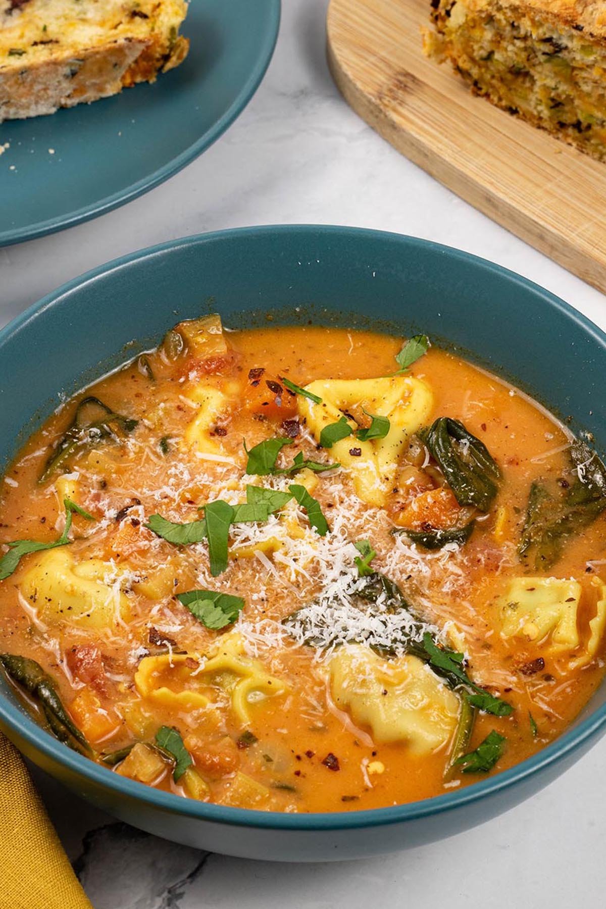 A portion of tomato and tortellini soup in a teal bowl with a plate of sliced leek and red leicester soda bread in the background.