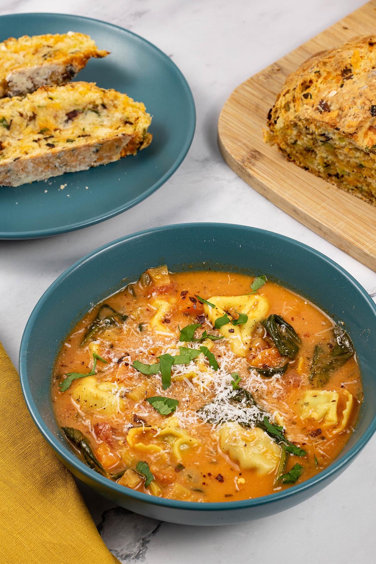 A portion of tomato and tortellini soup in a teal bowl with a plate of sliced leek and red leicester soda bread in the background.
