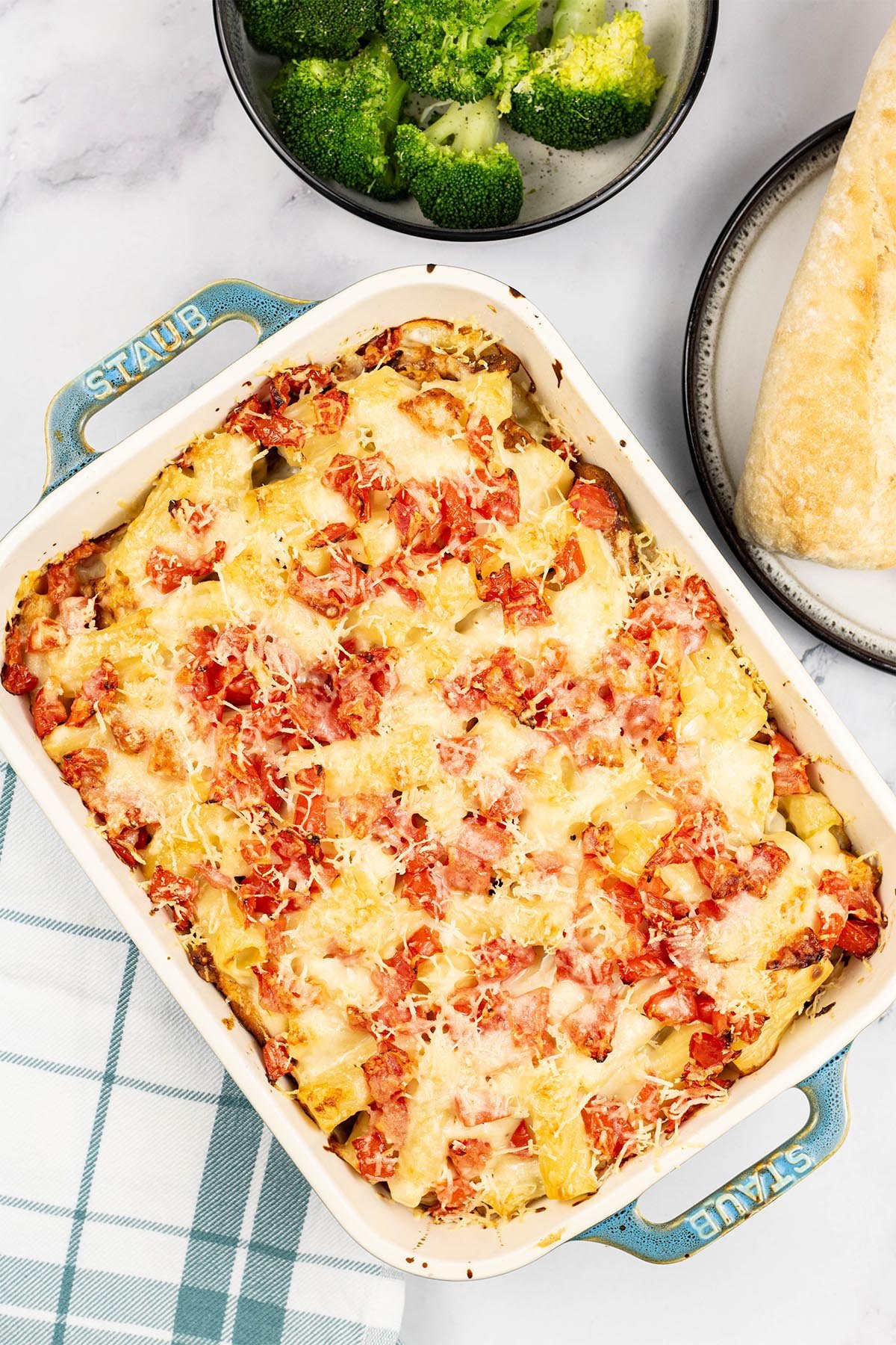 Chicken pasta bake in a rectangle Staub ovenproof dish with a bowl of broccoli and plate of crusty bread on the side.