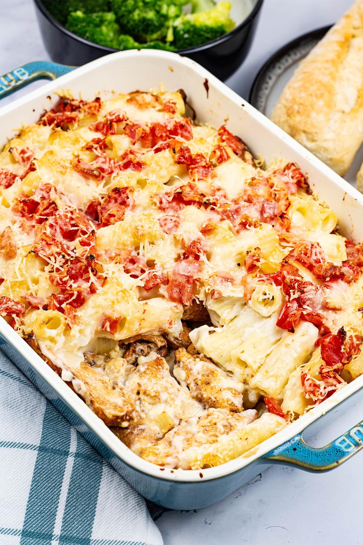 Chicken pasta bake in a rectangle Staub ovenproof dish with a bowl of broccoli and plate of crusty bread on the side.
