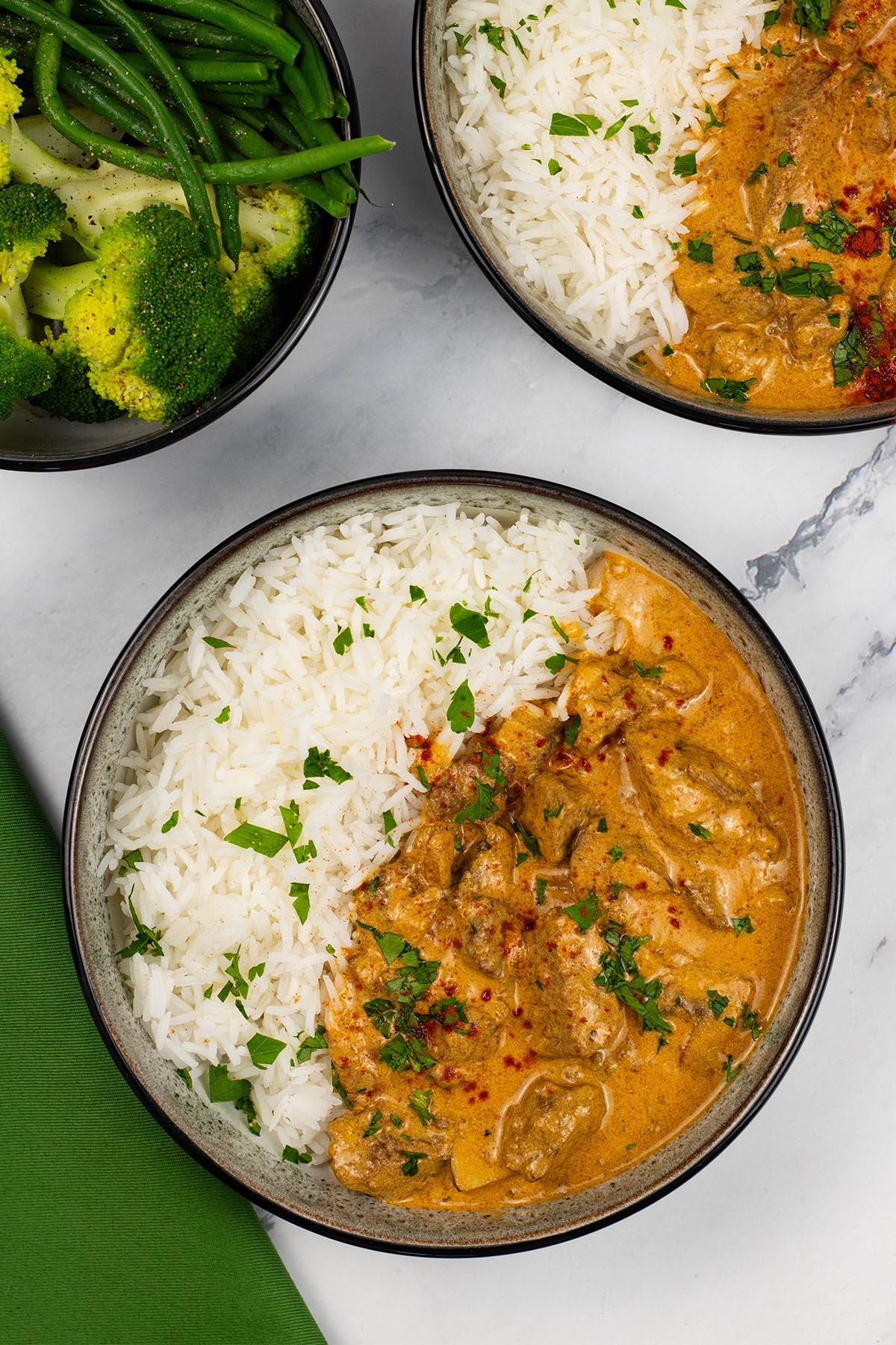 Two portions of slow cooker beef stroganoff in black and white bowls with rice and a bowl of green veg on the side.