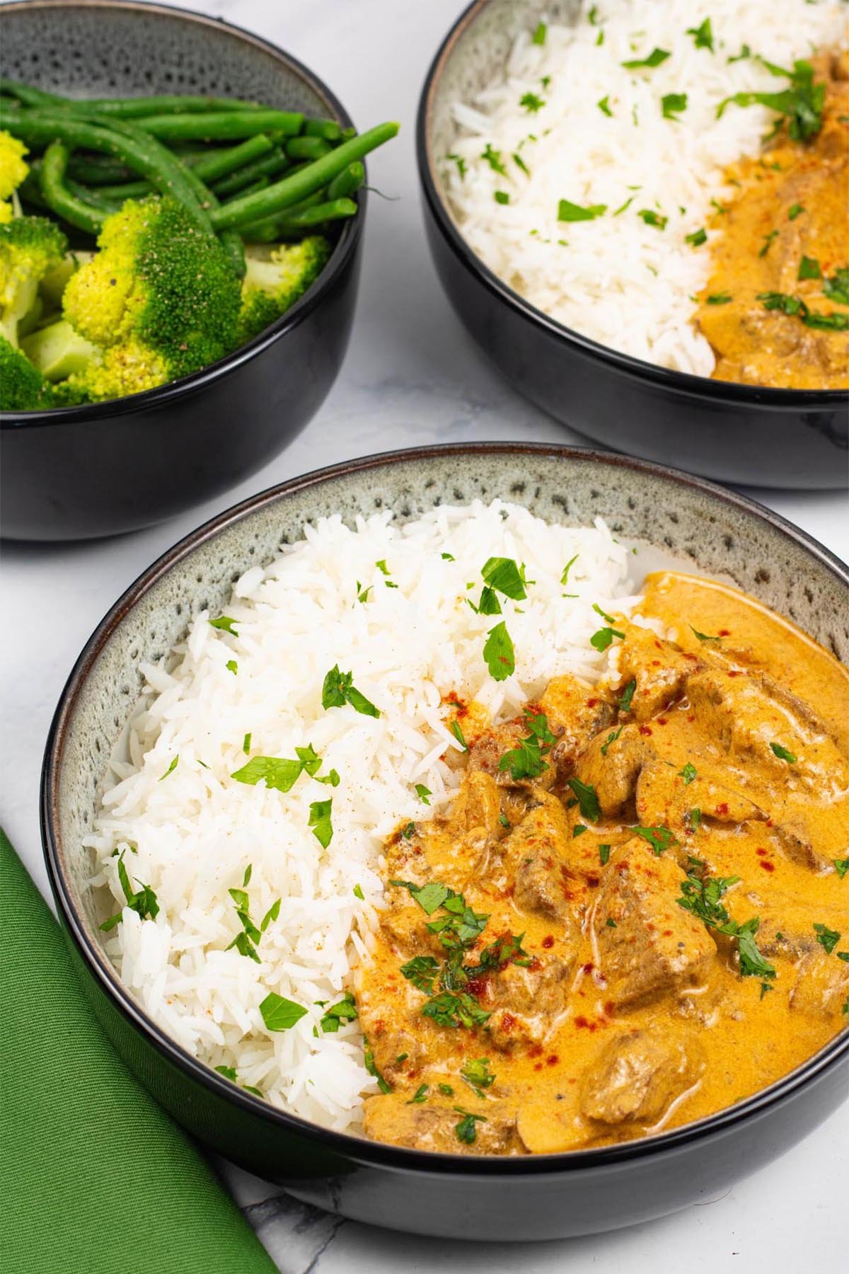 Two portions of slow cooker beef stroganoff in black and white bowls with rice and a bowl of green veg on the side.