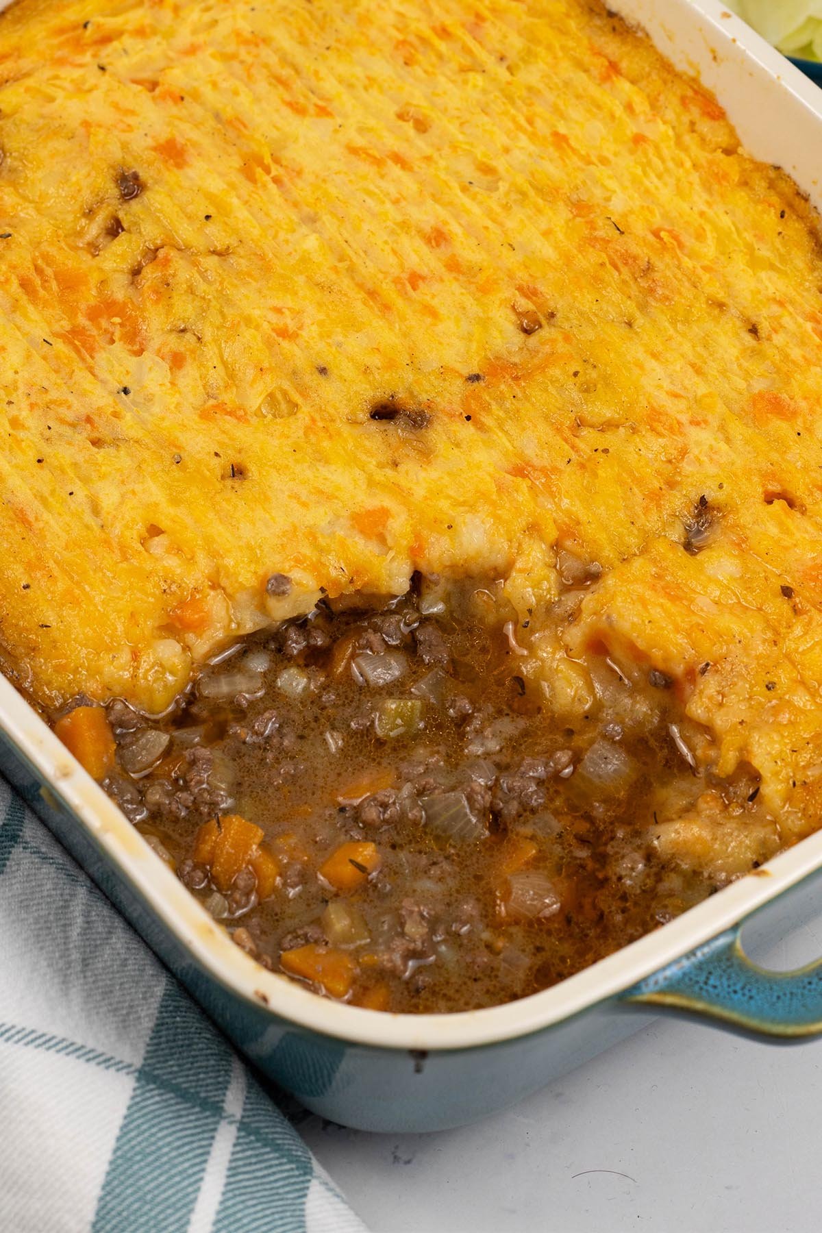 Close up of venison mince pie in a rectangular Staub ovenproof dish with a checked teal and white towel on the side.