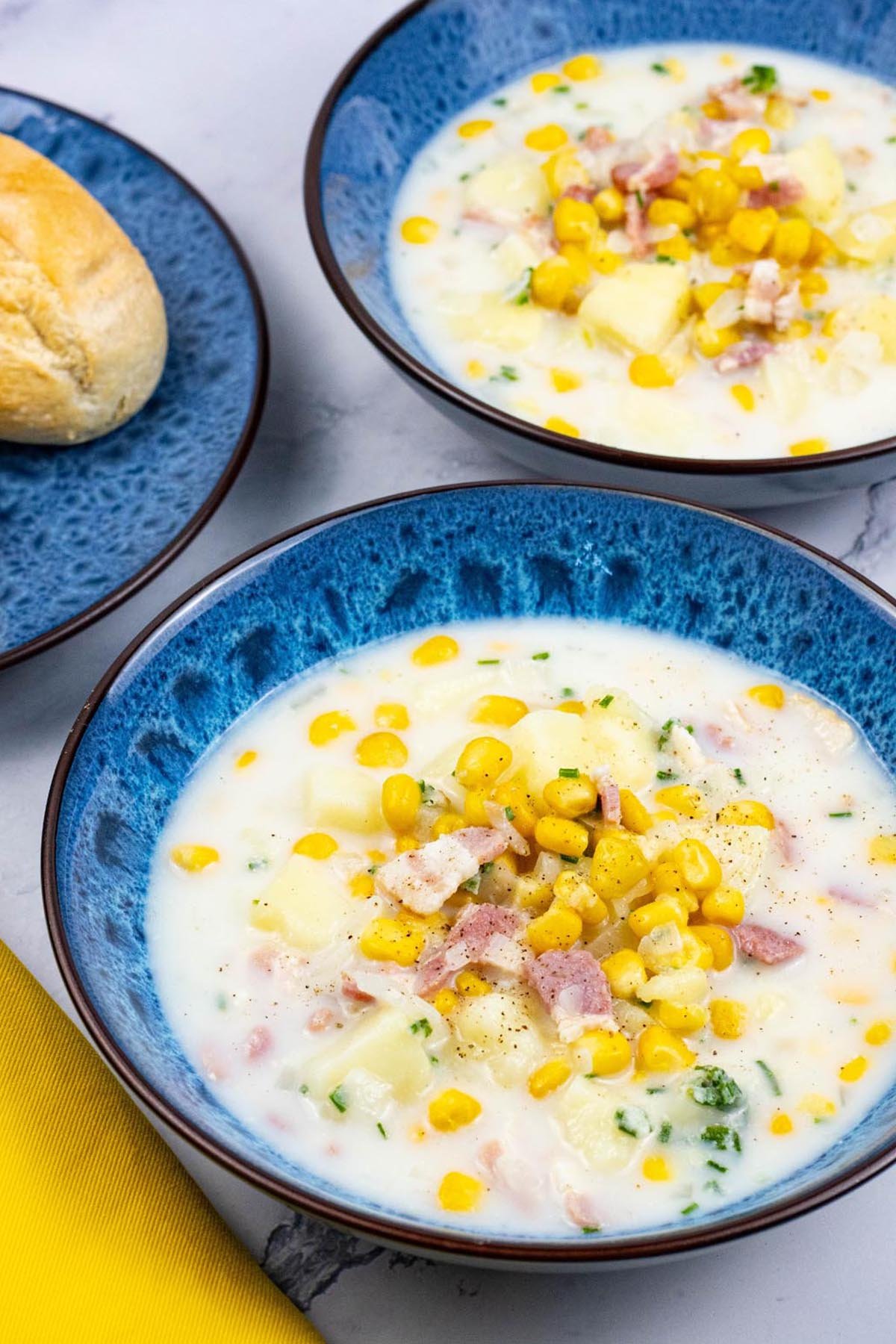 Two bowls of bacon and sweetcorn chowder in blue patterned bowls with a plate of crusty buns on the side.