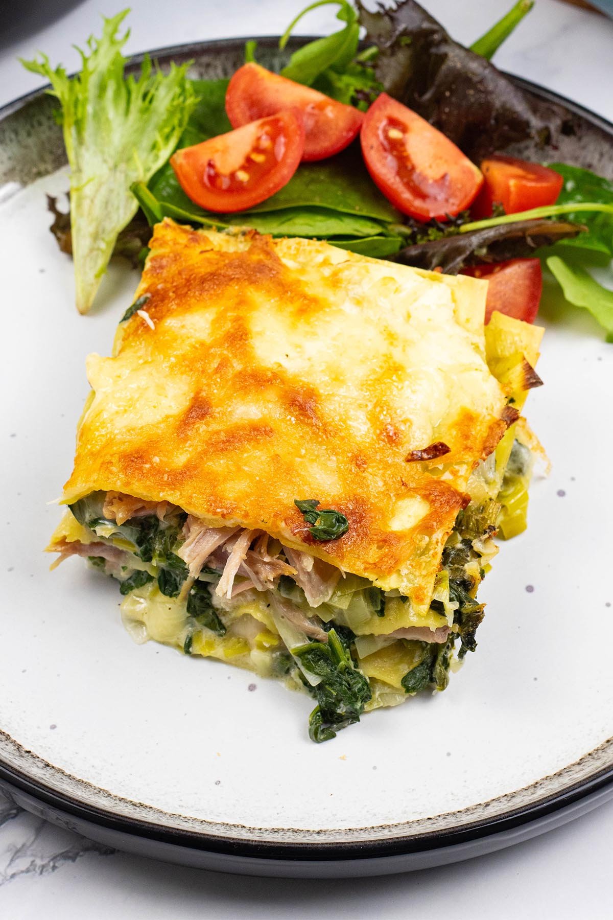A portion of ham, spinach and mozzarella lasagne on a black and white dinner plate with a green leaf and tomato salad.