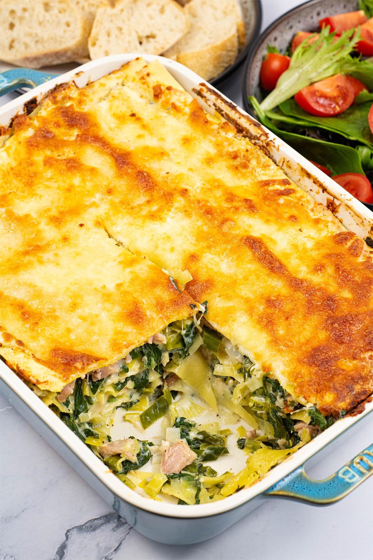 Ham, spinach and mozzarella lasagne in a rectangle Staub oven dish with a portion removed, a bowl of salad and plate of crusty bread on the side.