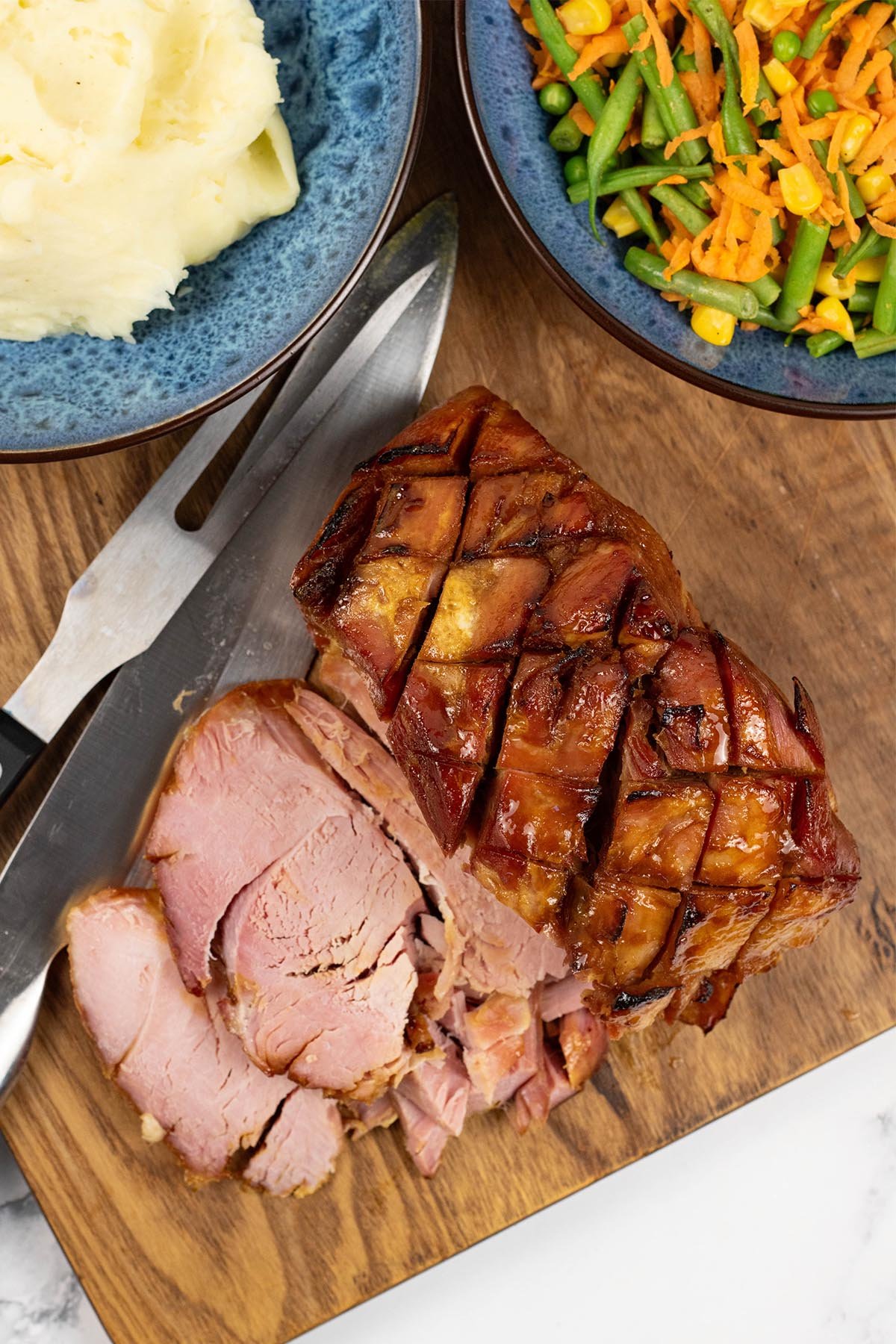 Slow cooker honey roast ham on a wooden board with two slices cut and bowls of mashed potatoes and warm carrot salad on the side.