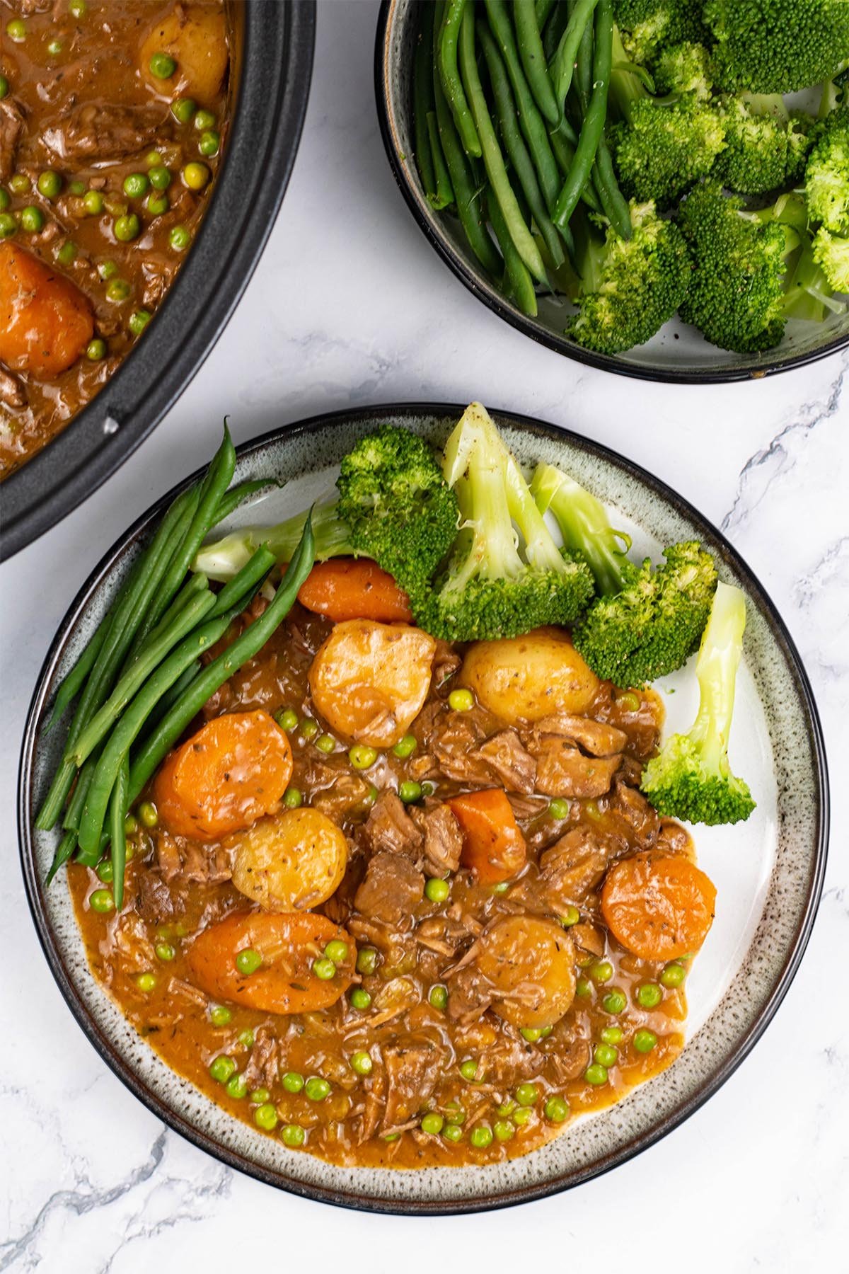 A portion of slow cooker old fashioned beef stew on a black and white patterned dinner plate with broccoli and green beans on the side.