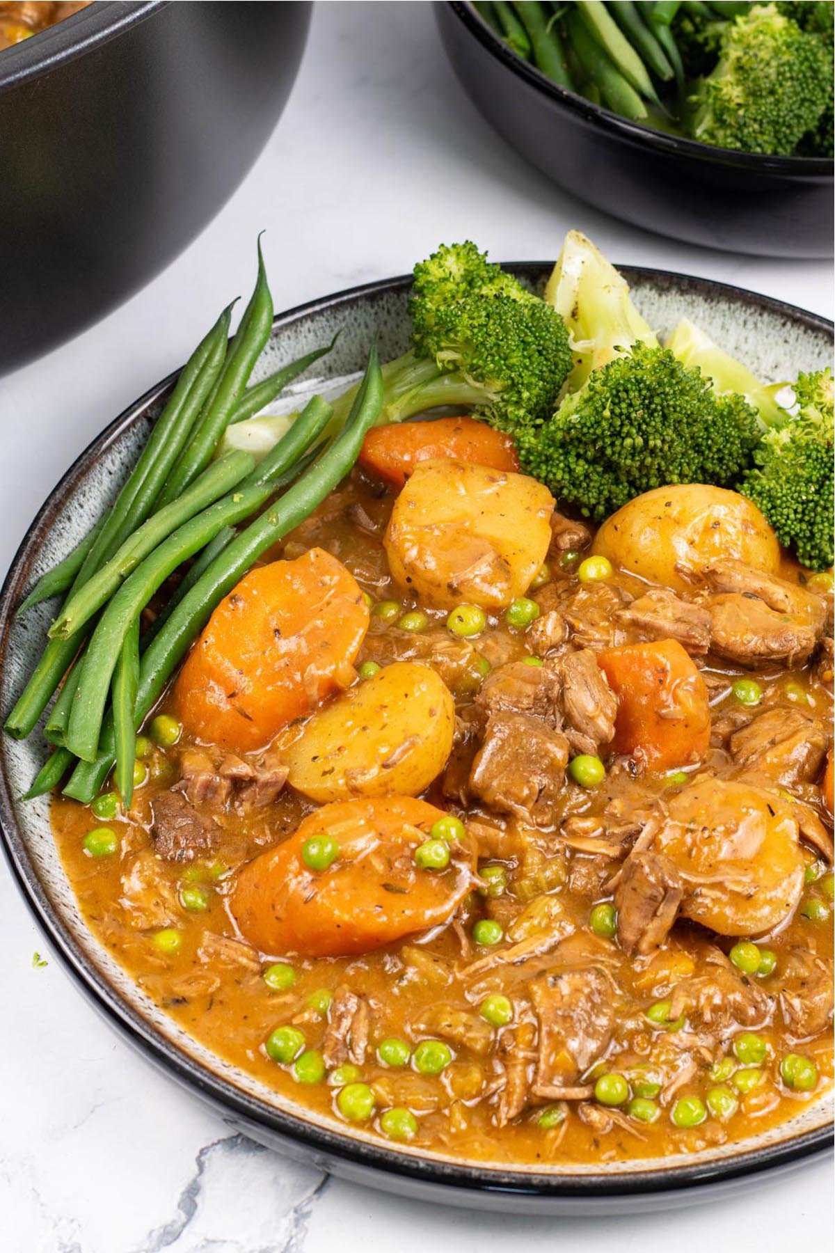 A portion of slow cooker old fashioned beef stew on a black and white patterned dinner plate with broccoli and green beans on the side.