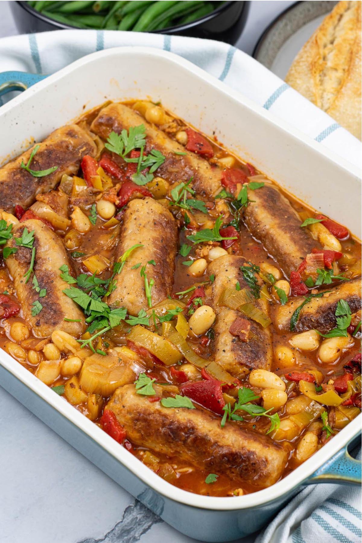 Slow cooker sausage casserole in a rectangle Staub serving dish with baguette and green beans in the background.