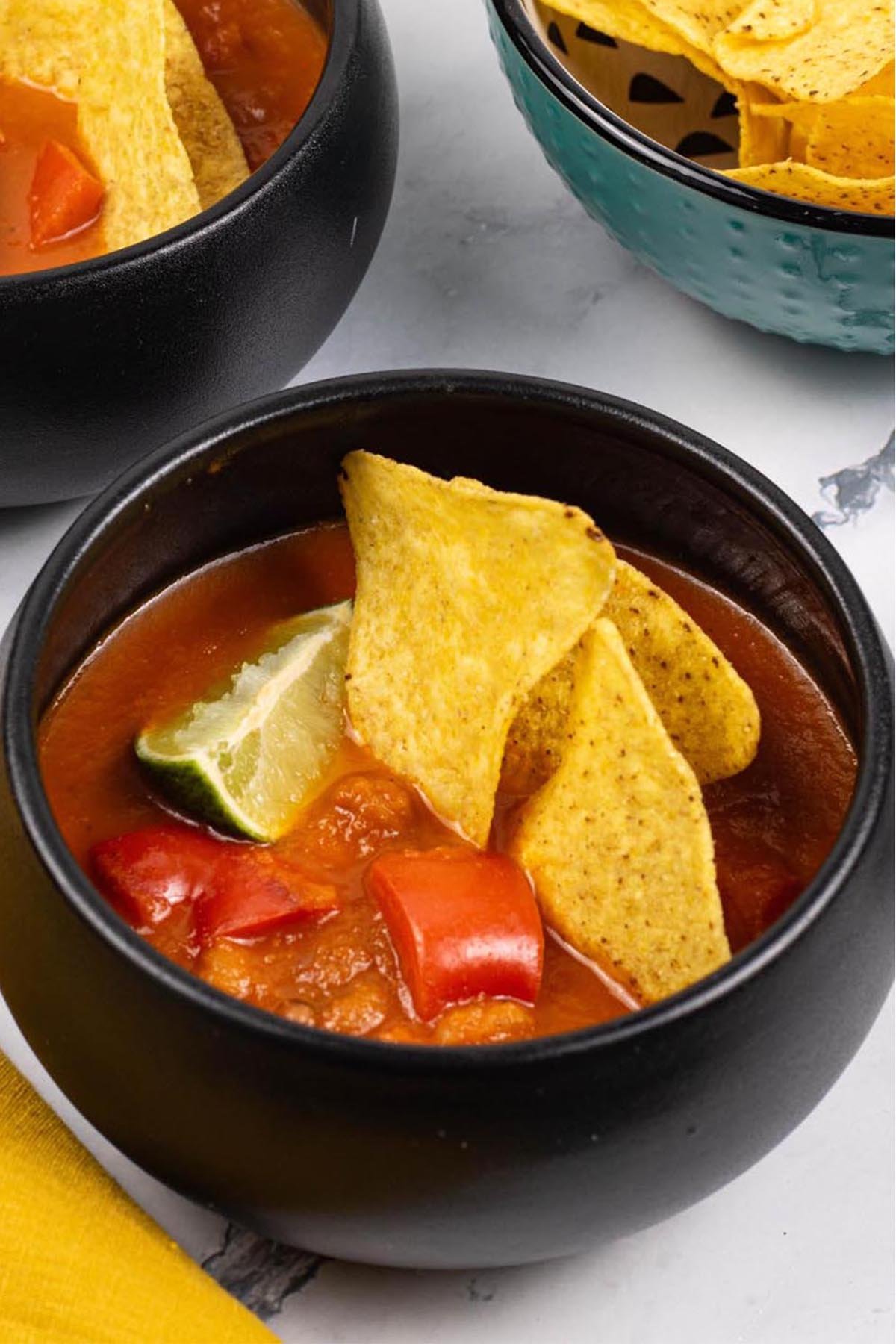 Two portions of spicy chilli bean soup in black bowls garnished with tortilla crisps and a wedge of lime.