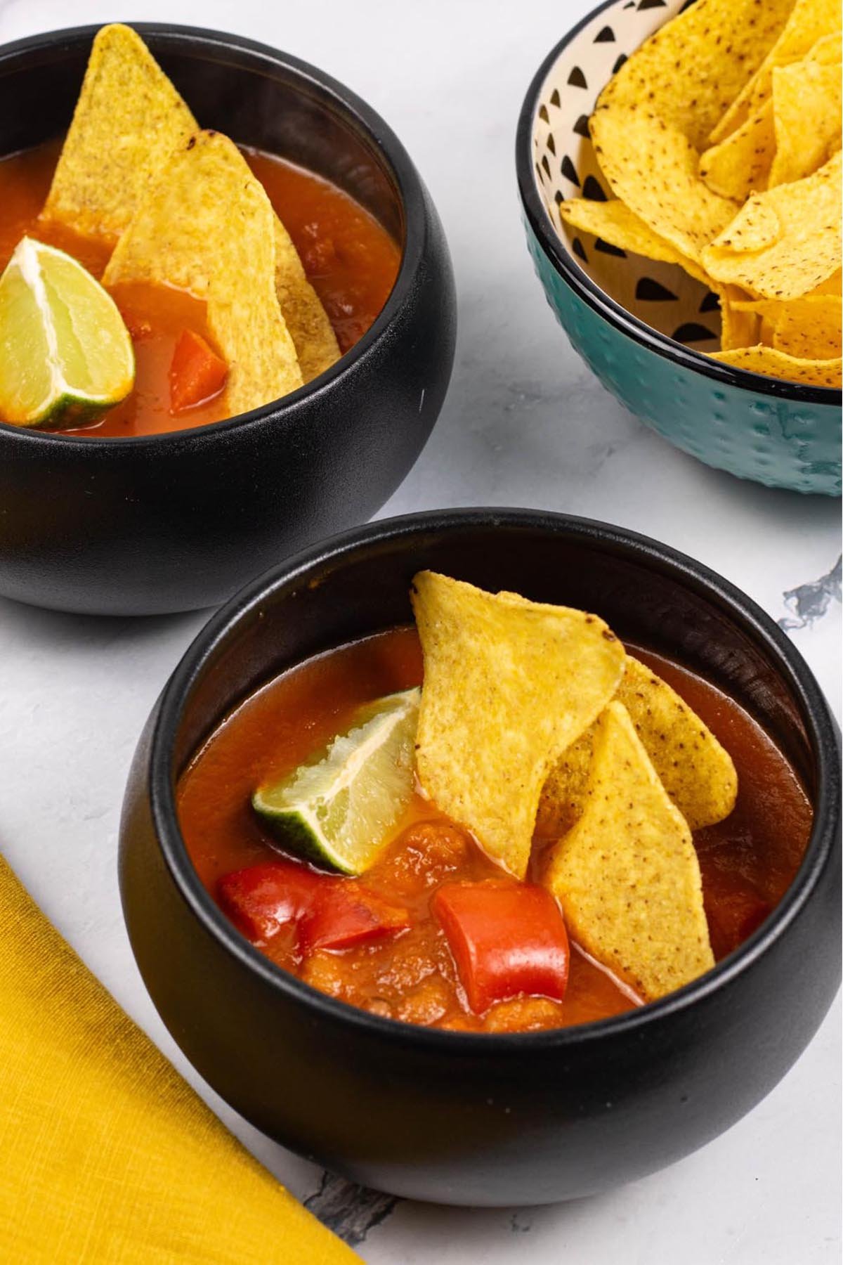 Two portions of spicy chilli bean soup in black bowls garnished with tortilla crisps and a wedge of lime.