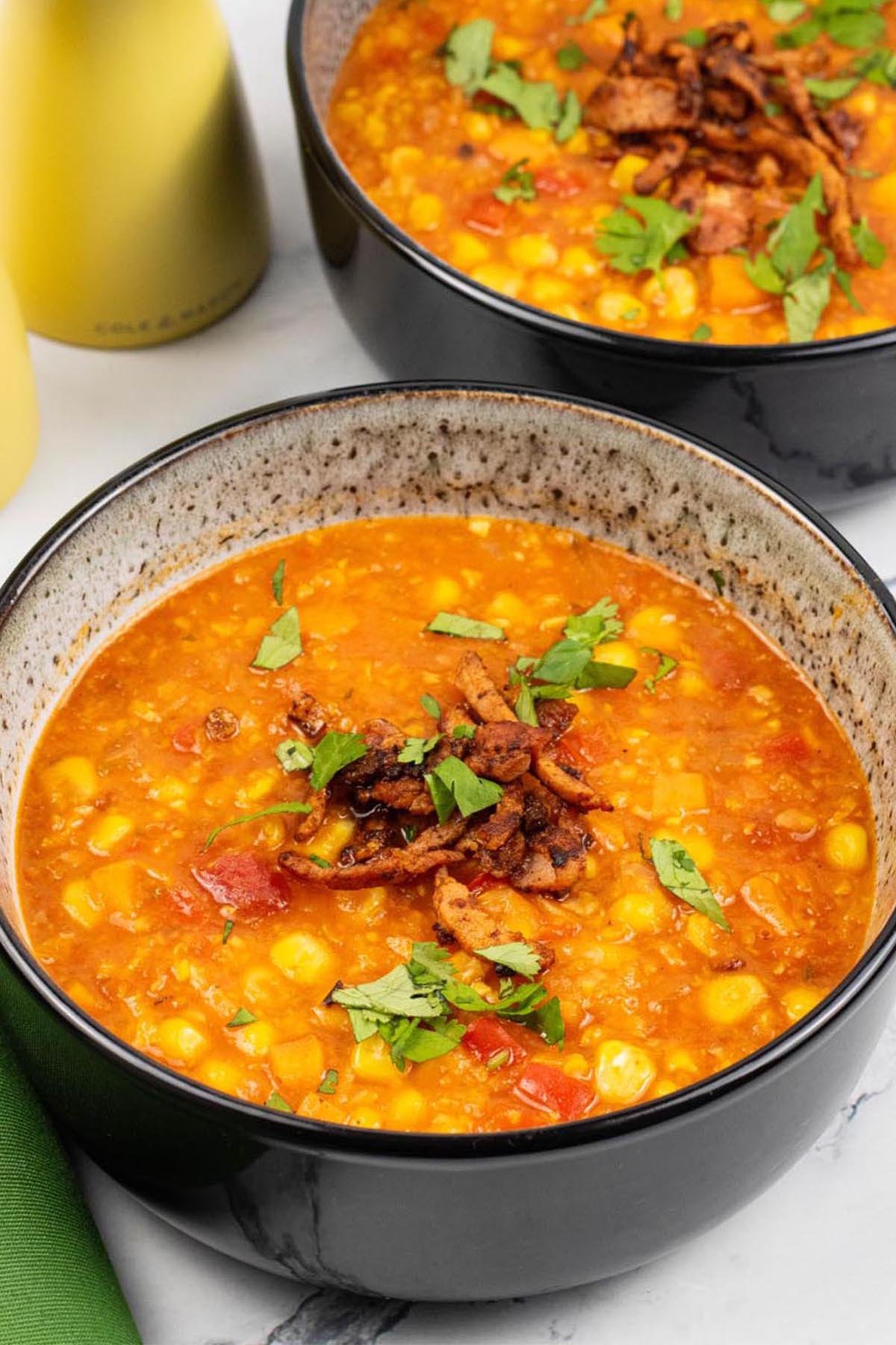 Two portions of spicy sweetcorn soup in black patterned bowls with yellow salt and pepper grinders and a green napkin on the side.