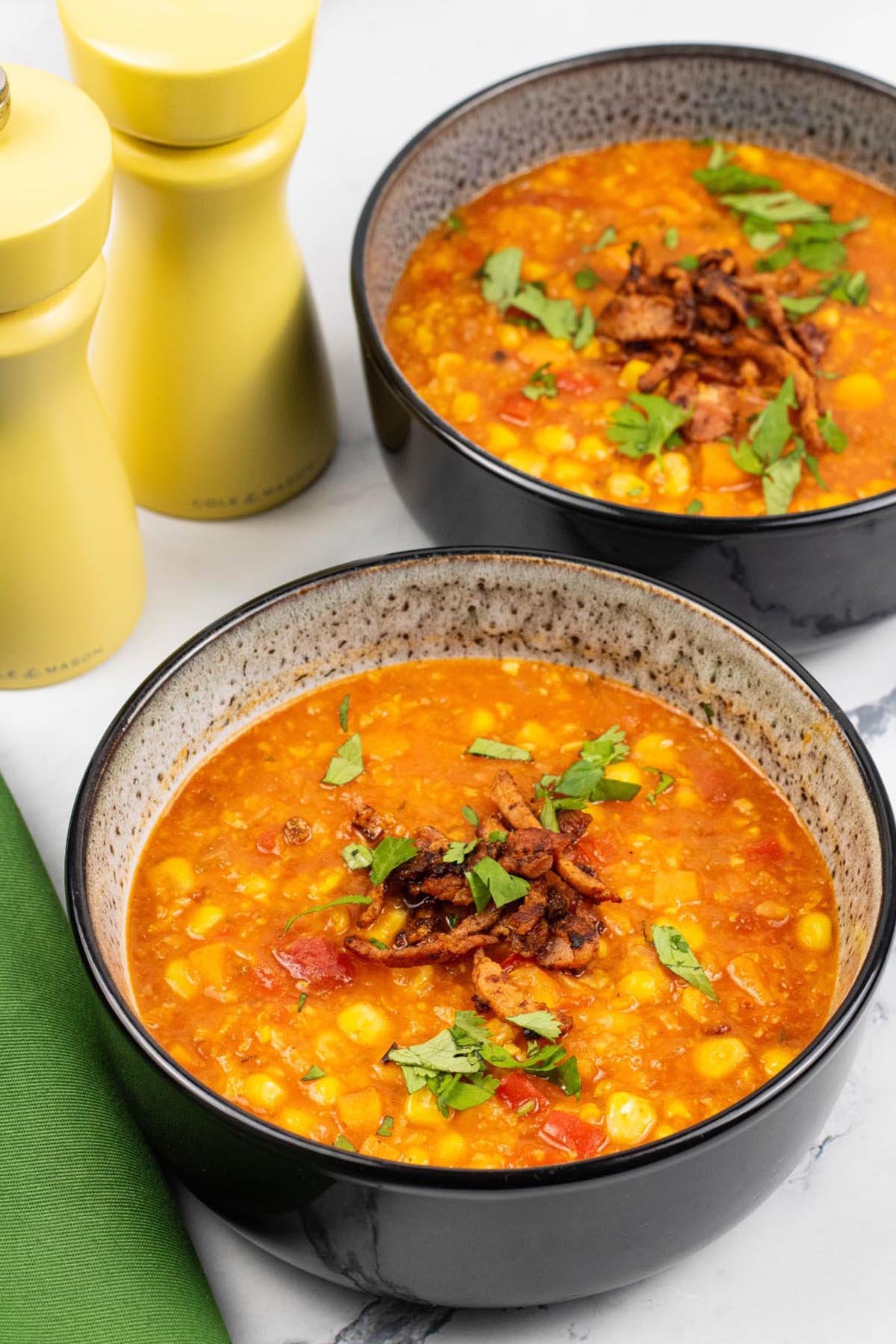 Two portions of spicy sweetcorn soup in black patterned bowls with yellow salt and pepper grinders and a green napkin on the side.