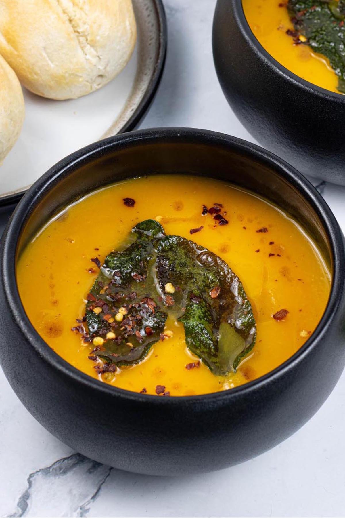 Two portions of squash and carrot soup with crispy sage leaves in black bowls with a plate of crusty rolls on the side.
