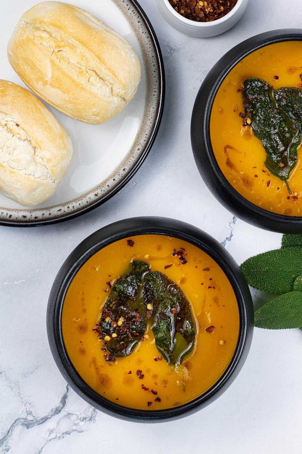 Two portions of squash and carrot soup with crispy sage leaves in black bowls with a plate of crusty rolls on the side.